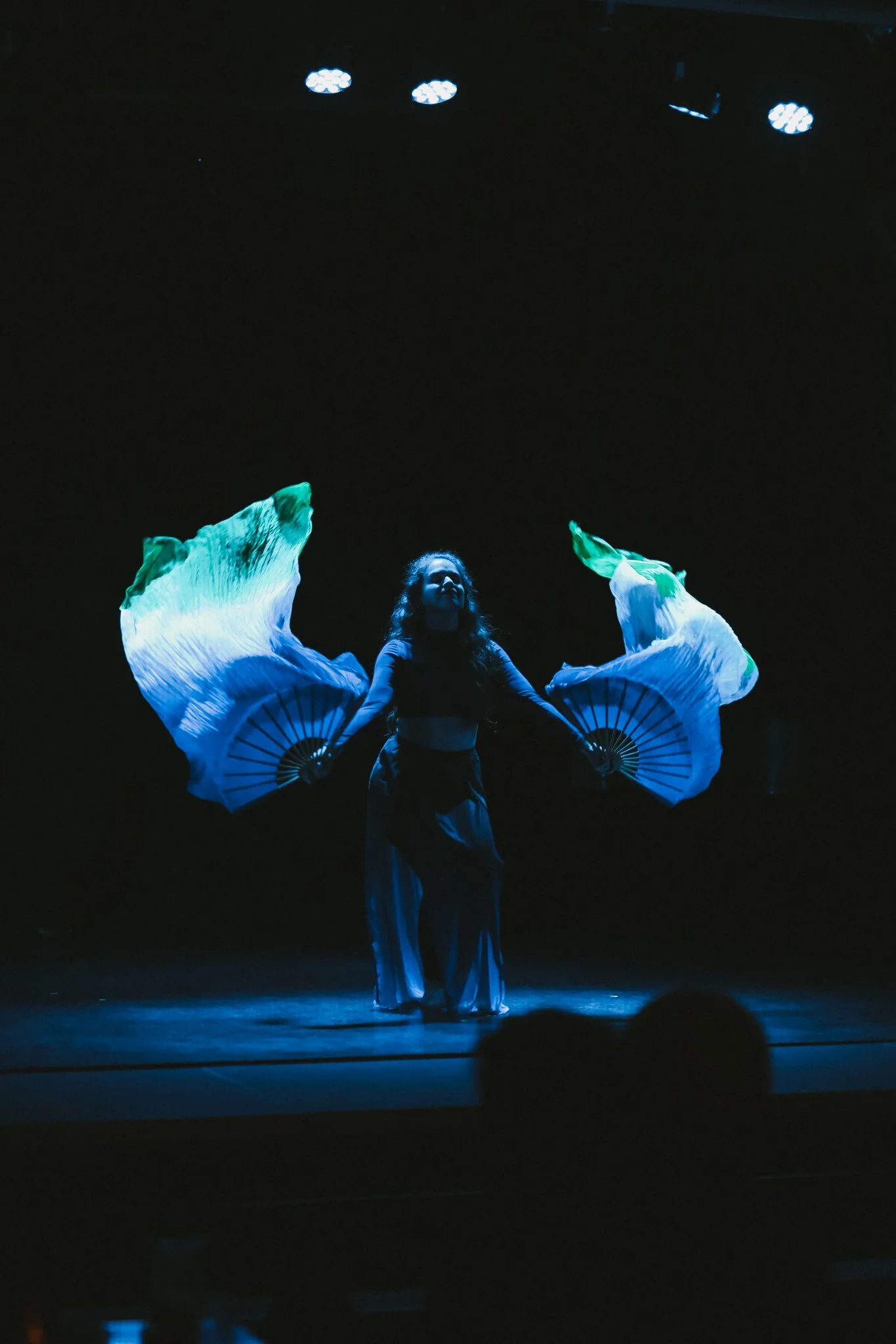 A female performer holding two large, illuminated fans on a dark stage.