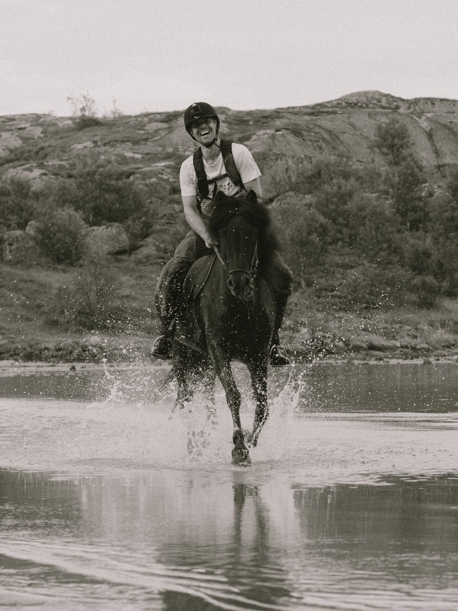A man riding a horse through a shallow body of water, smiling, with a mountainous landscape in the background, in black and white.
