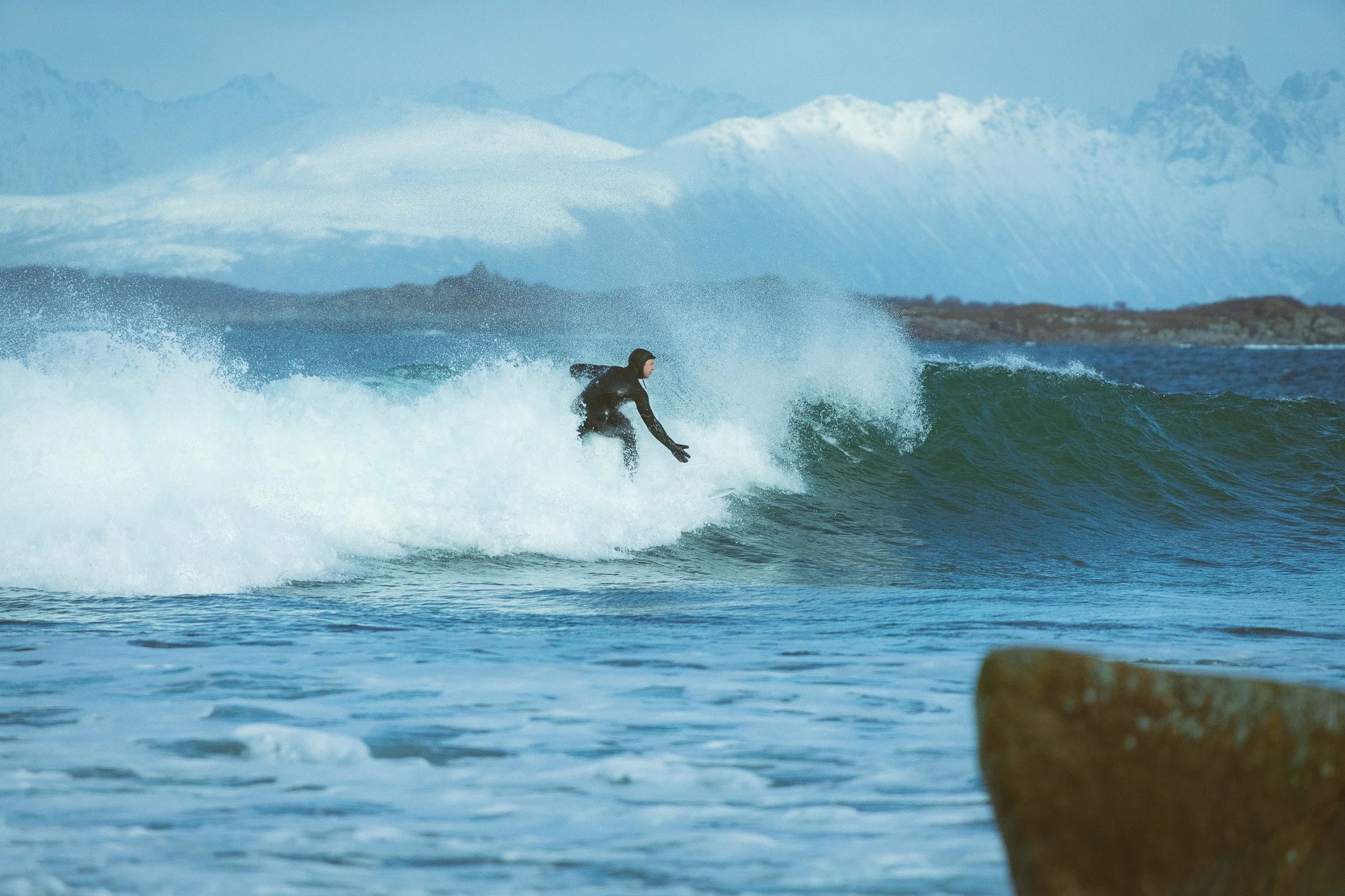 A person in a wetsuit surfing on a wave with snow-covered mountains in the background.
