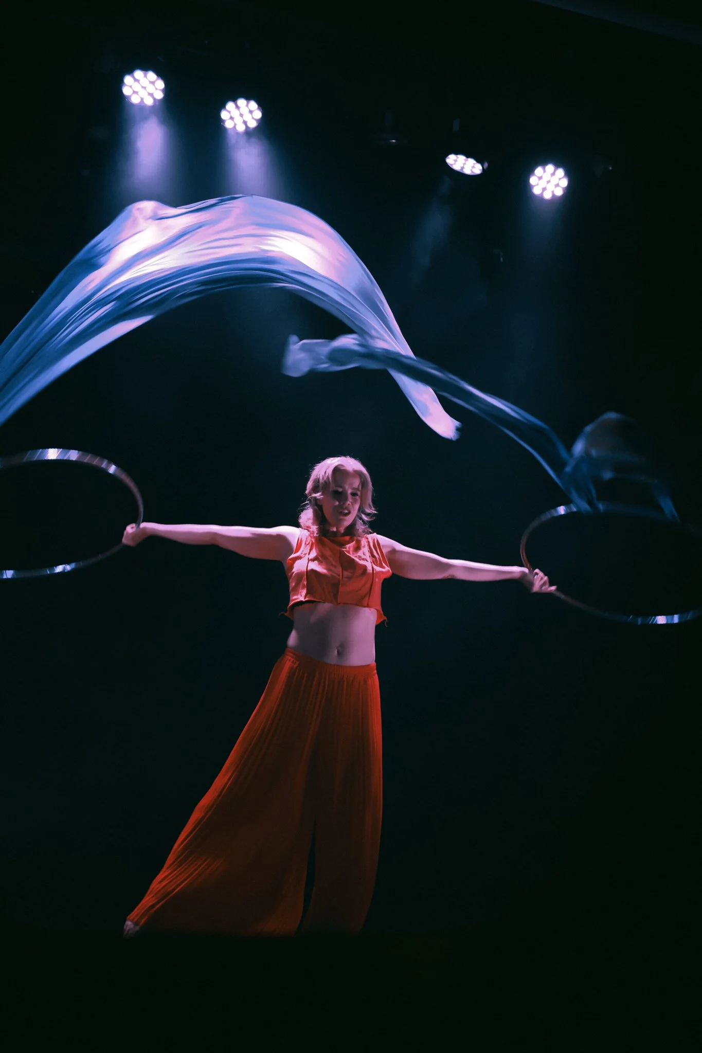 A performer in an orange outfit balancing on stage while spinning two hula hoops with blue fabric flowing above her.