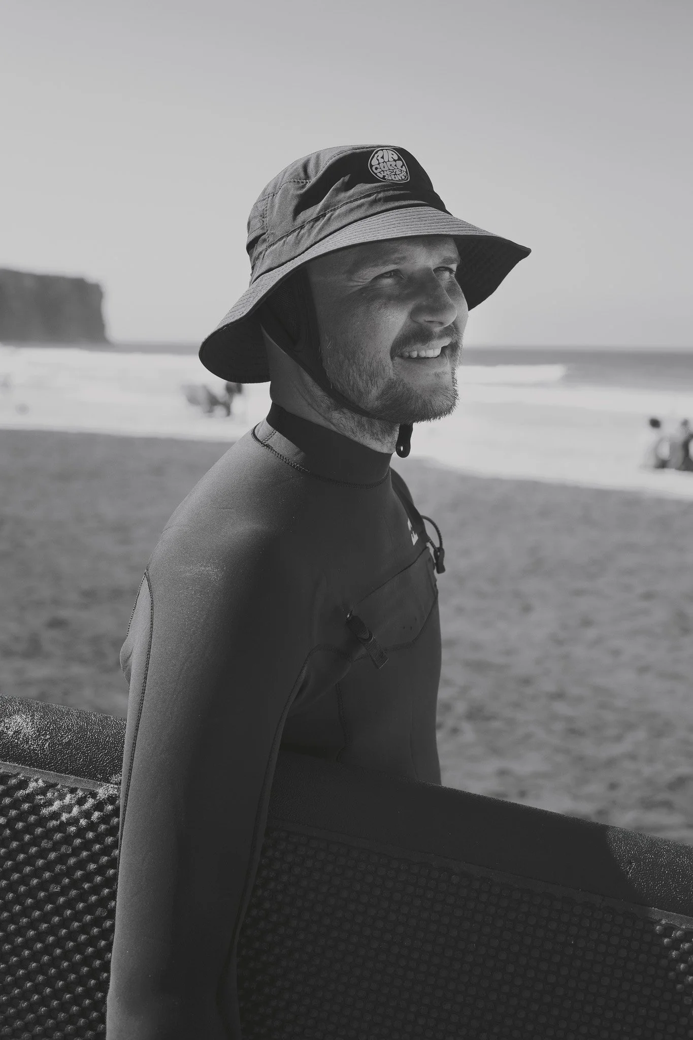 A man wearing a bucket hat, a wetsuit, and a life jacket standing on a beach with the ocean and cliffs in the background.