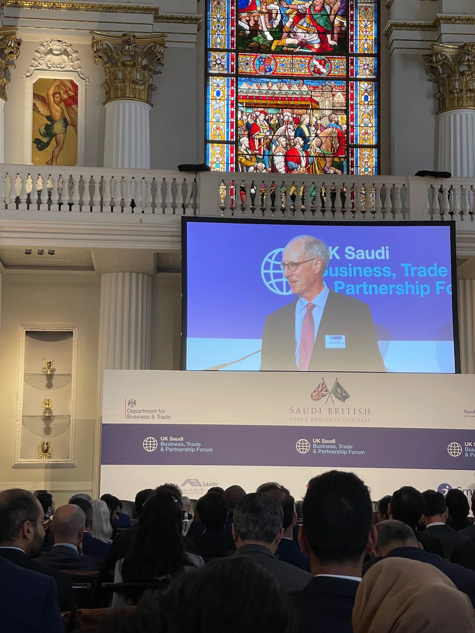 A man is giving a speech at a conference titled 'UK Saudi Business, Trade & Partnership Forum,' with a large audience seated in front. The backdrop displays the logos of the Department for Business & Trade and the Saudi British Joint Business Council, along with flags of the UK and Saudi Arabia. Behind the speaker, a stained glass window depicts a historical scene with multiple figures, and the room has ornate columns and religious artwork.