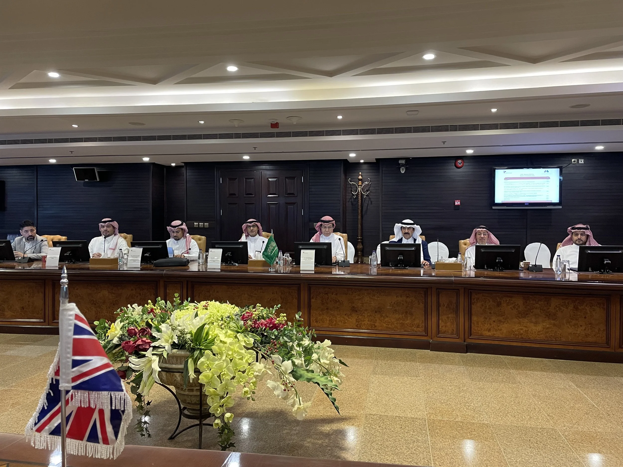 A formal meeting with nine men sitting at a long wooden table with computer screens, in a conference room decorated with flowers and small flags, including the United Kingdom flag in the foreground, and Saudi Arabian flags on the table. The men are wearing traditional Middle Eastern attire, including thobes and shemagh head coverings.