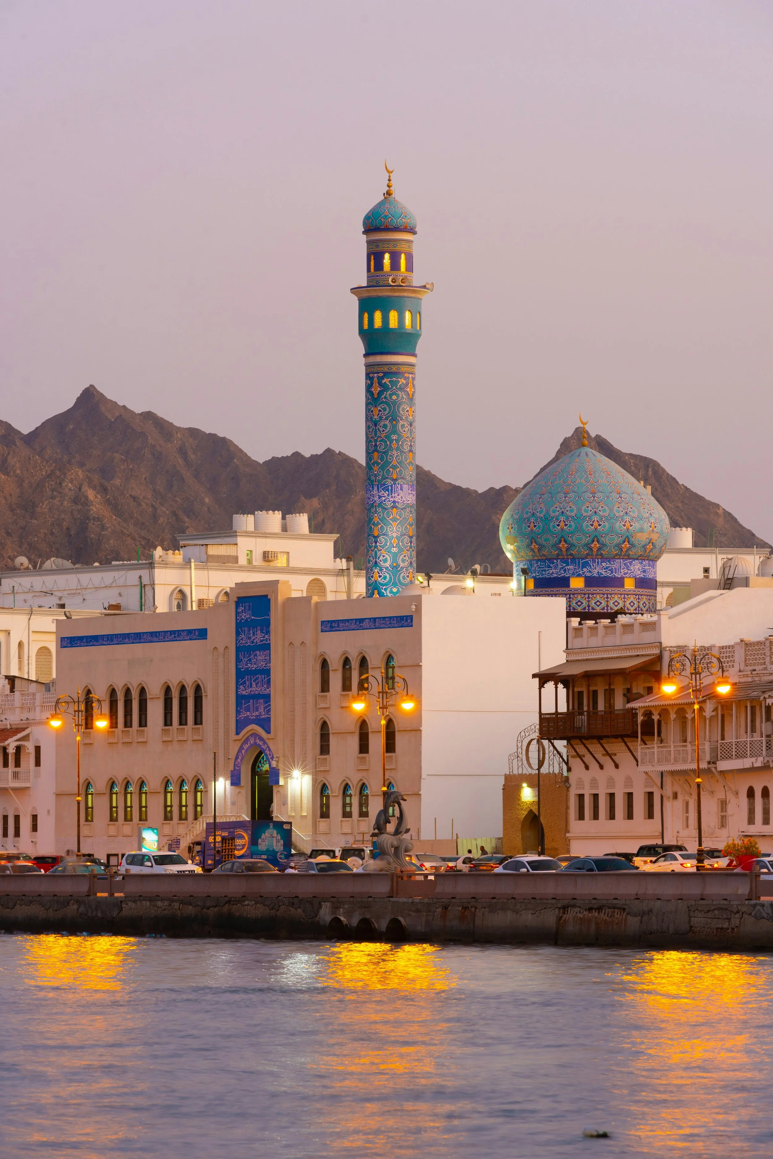 A mosque with a tall, decorated minaret and blue domed roof, set against a mountainous background and reflecting in the water in the foreground.