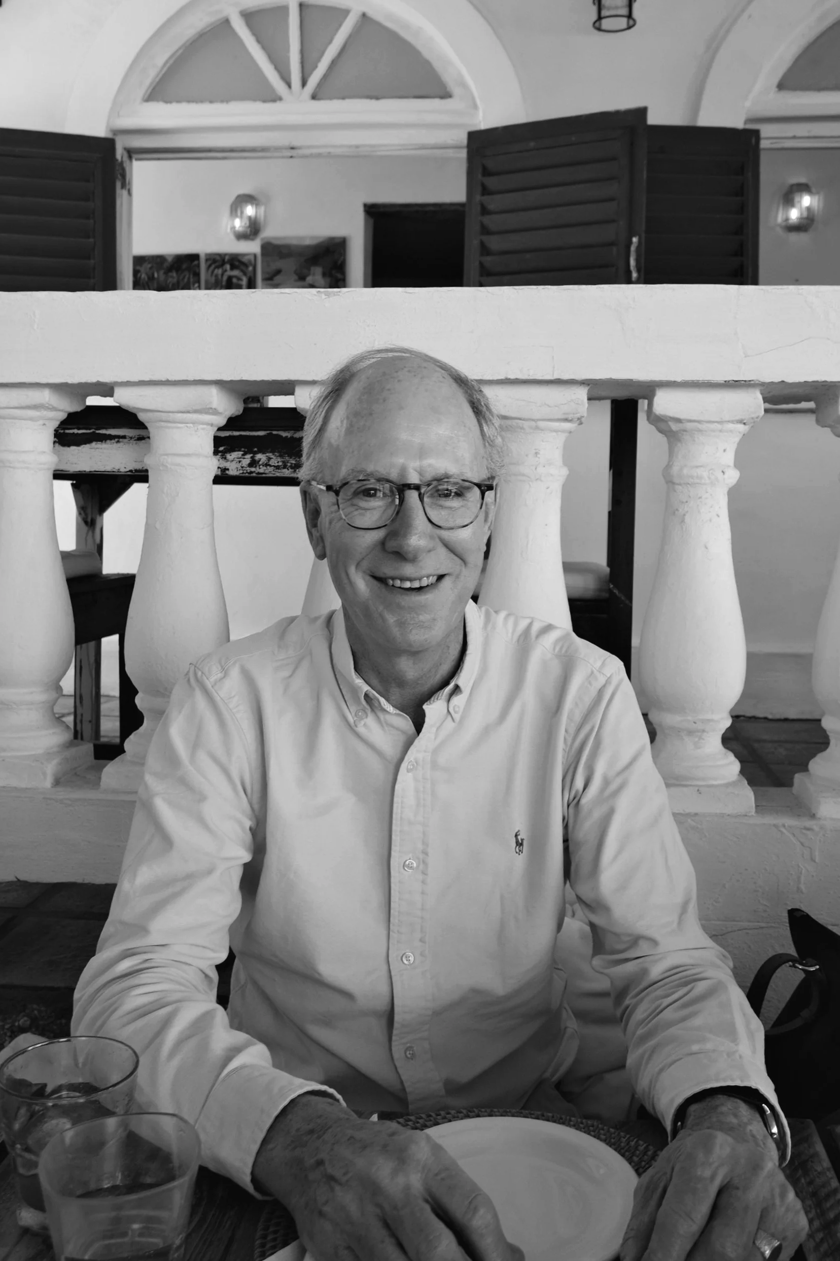 A smiling man with glasses wearing a light-colored button-up shirt sitting at a table inside a building with arched windows and shutters in the background.