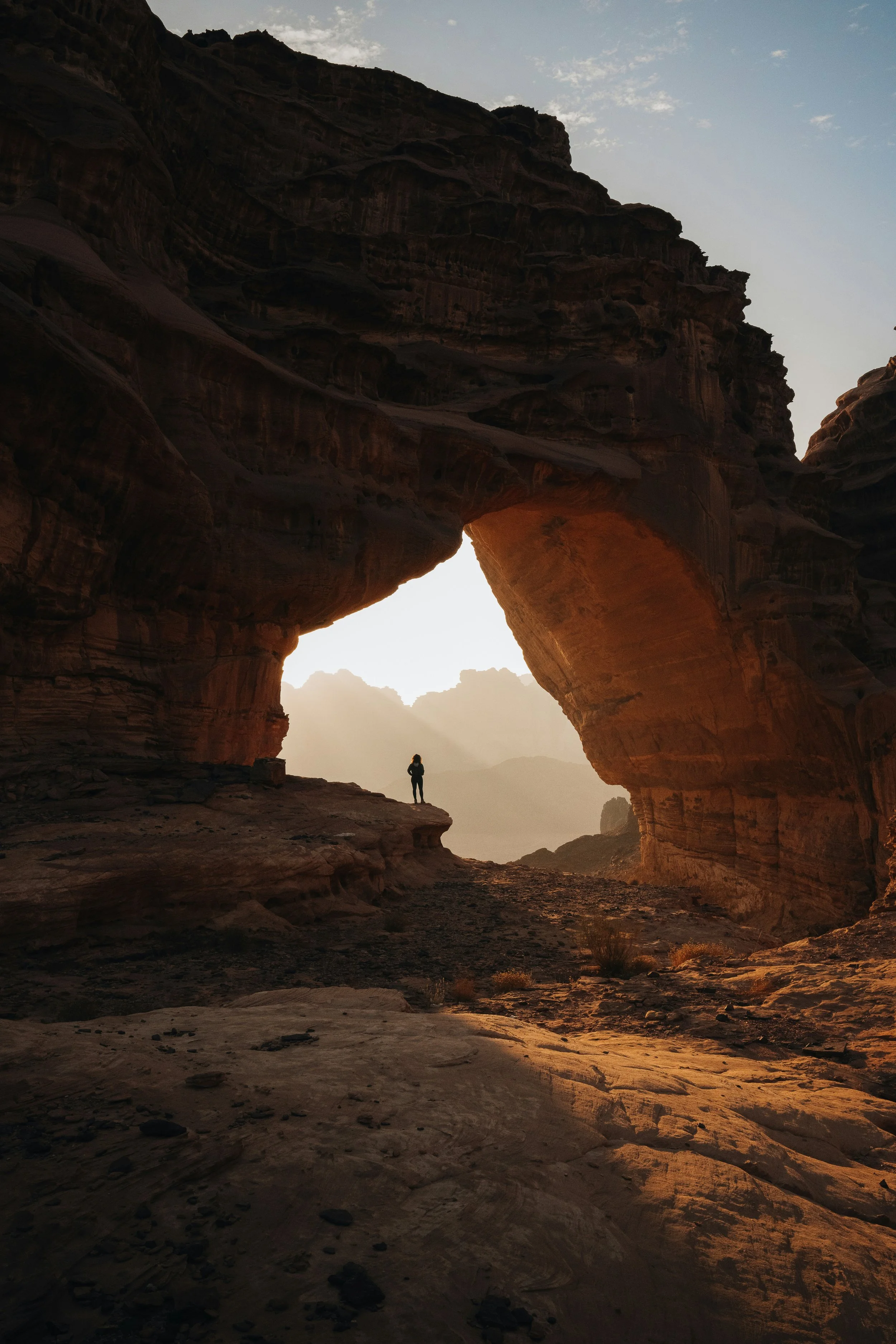 Silhouette of a person standing inside a large natural rock arch formation at sunset or sunrise, with mountains in the background and warm light illuminating the scene.