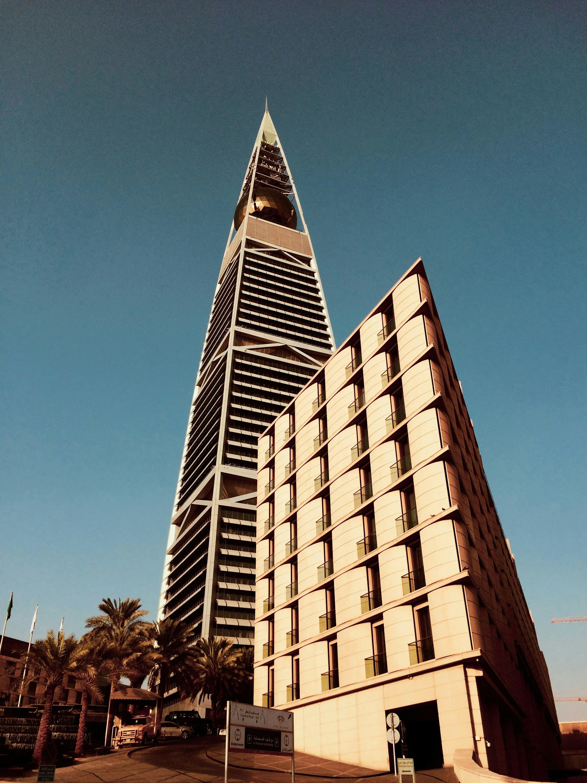 Low-angle view of the Salesforce Tower and a smaller modern building in San Francisco with palm trees in the foreground against a clear blue sky.