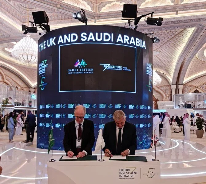 Two men in suits signing documents at a conference table with flags of the UK and Saudi Arabia, in front of a large digital display screen reading 'The UK and Saudi Arabia' at a business event in a luxurious venue.