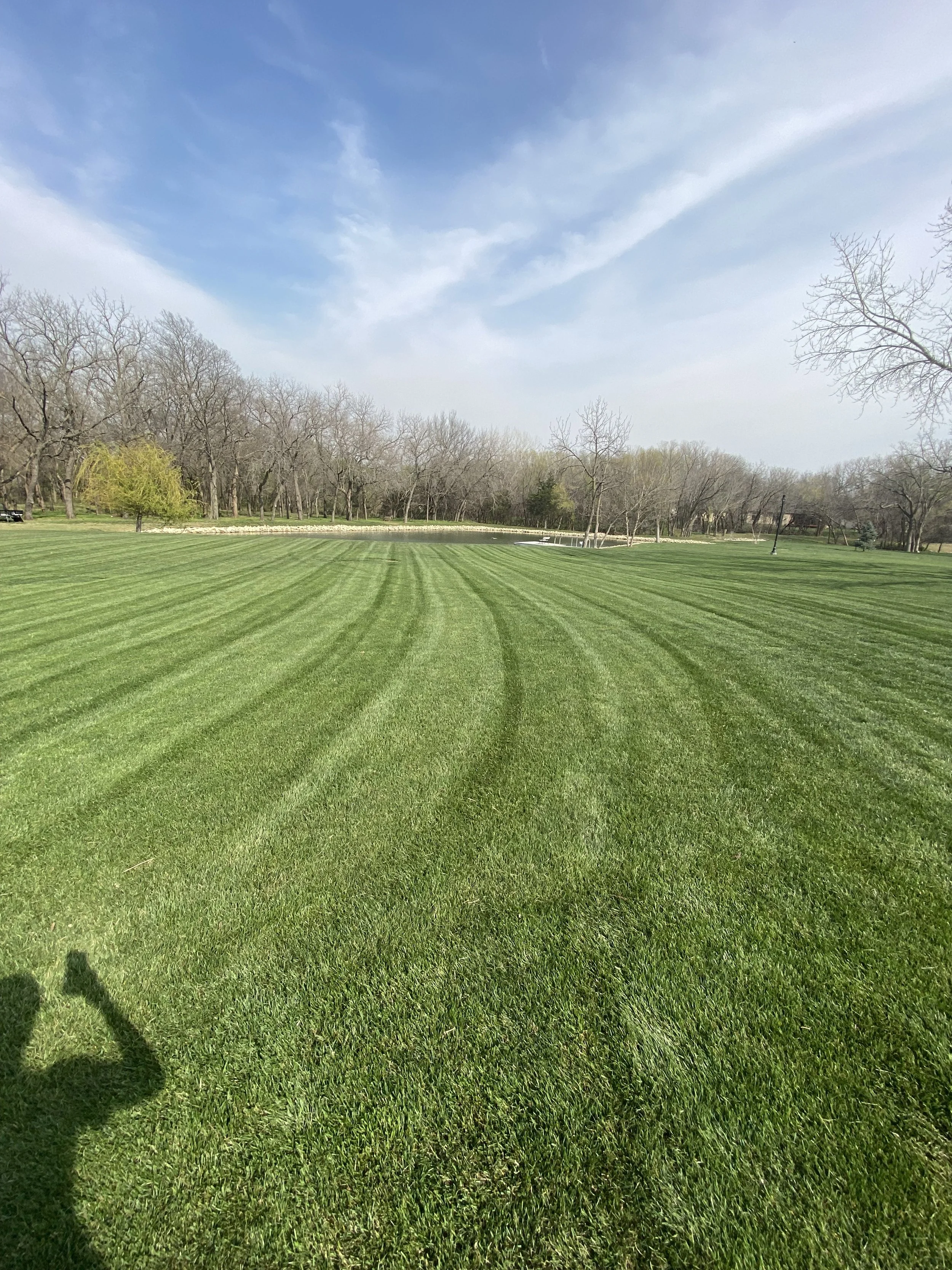 A large, well-maintained grassy field with tire tracks, bordered by trees with sparse leaves under a partly cloudy sky.