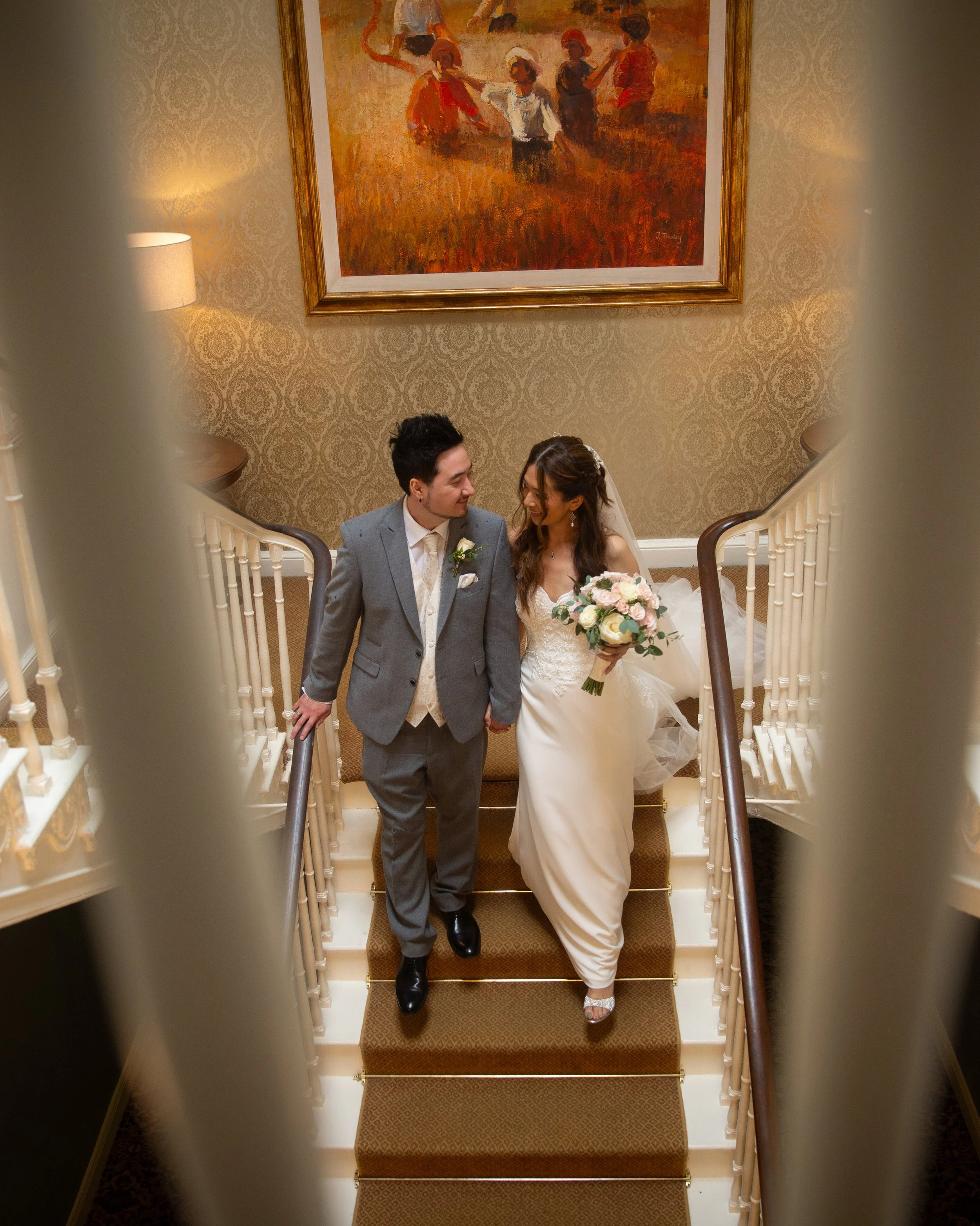 A bride and groom walking down a staircase, looking at each other and holding hands. The bride is holding a bouquet of flowers. The photo is taken from behind a railing, with a painting of children on a field hanging on the wall behind them.