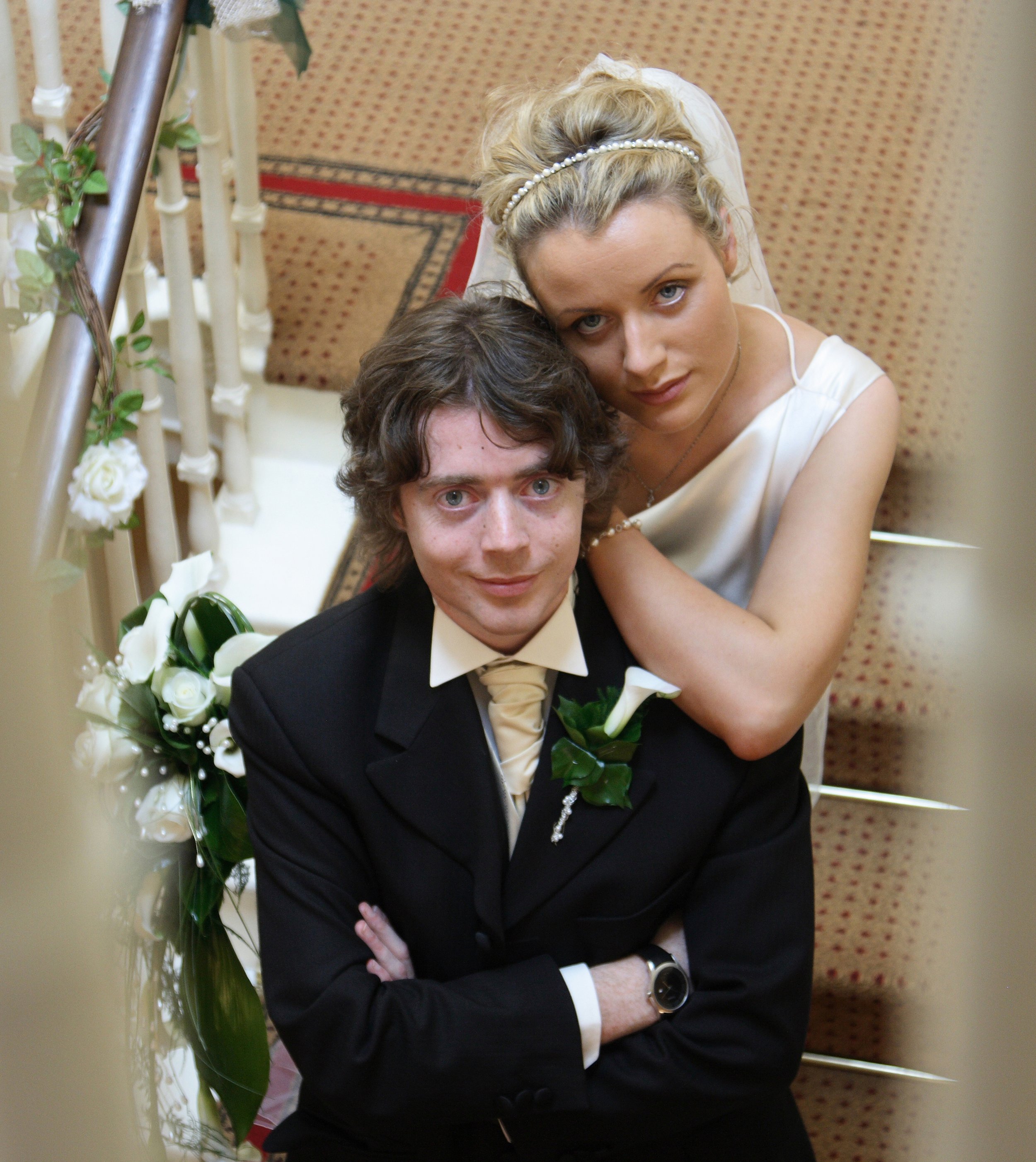 A bride with blonde hair in an updo wearing a white dress and a pearl headband leans over a groom with curly brown hair dressed in a black suit and cream tie, both posing on a staircase decorated with white roses and greenery, during a wedding celebration.