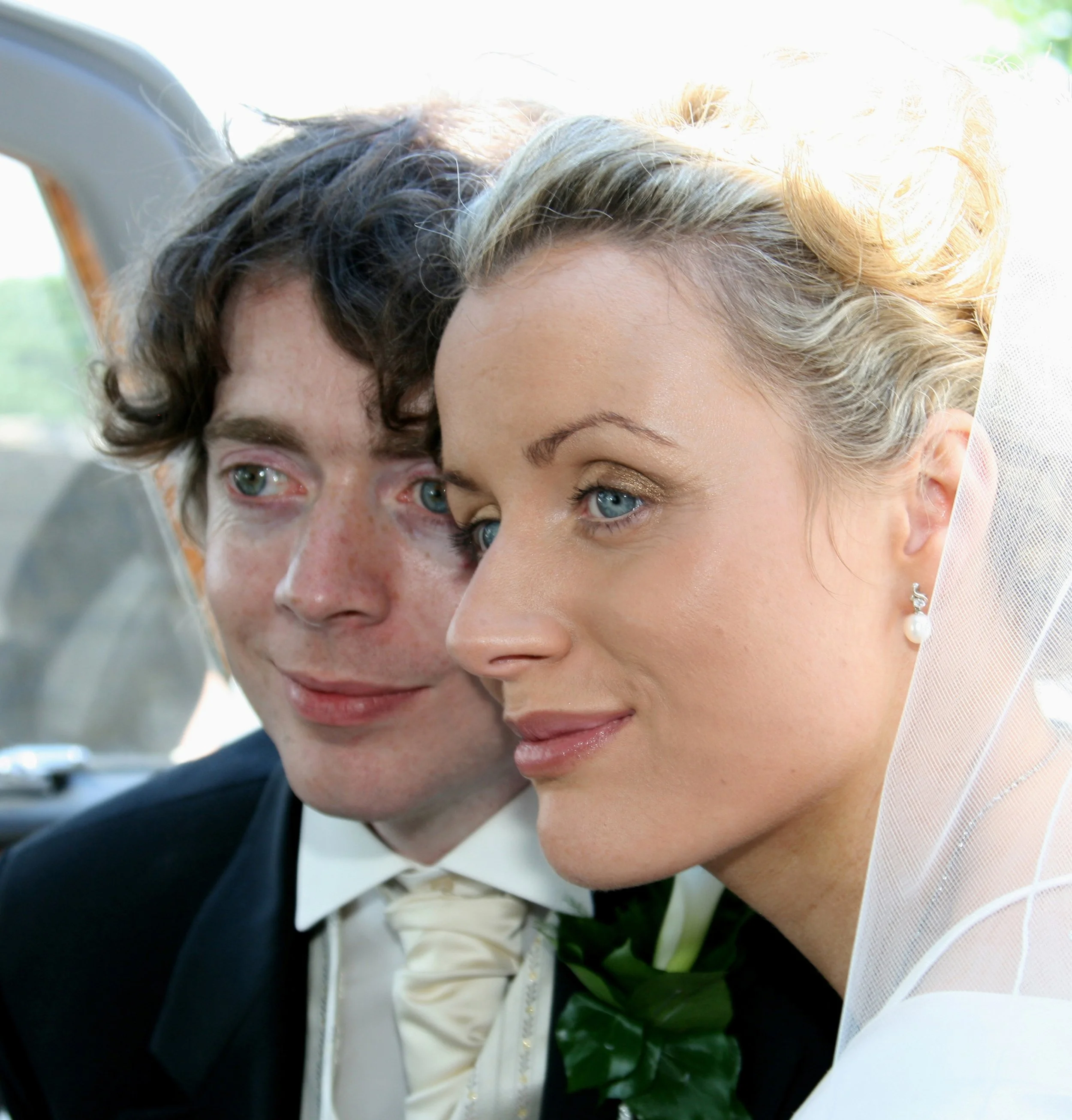 A bride and groom sitting close together during their wedding, inside a vehicle, with the bride in a white dress and veil, and the groom in a suit with a boutonnière.
