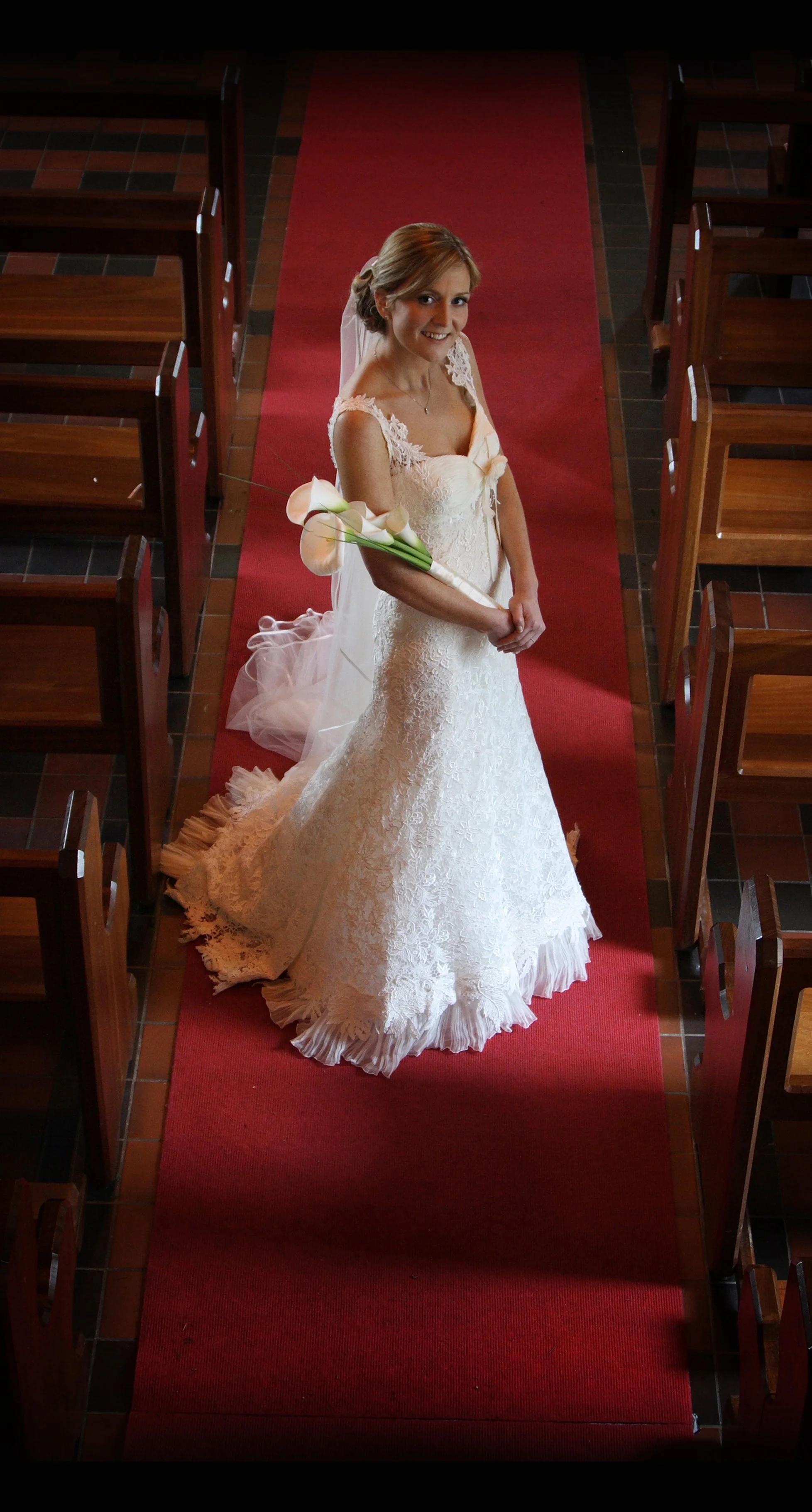 A bride in a wedding dress standing in the aisle of a church, holding a bouquet of calla lilies and smiling at the camera.