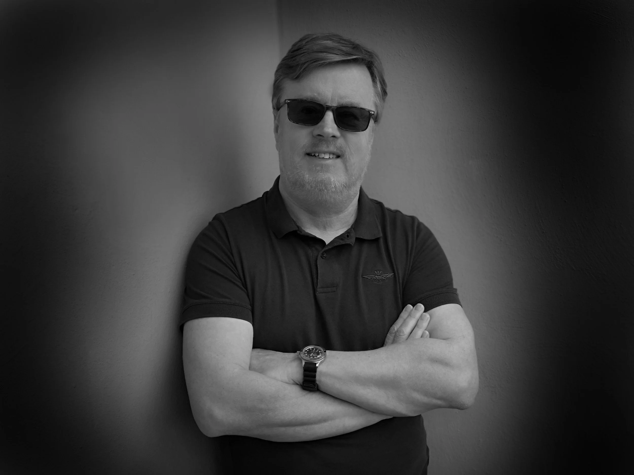 A man with sunglasses, a beard, and short hair standing against a plain wall with arms crossed, wearing a polo shirt and a wristwatch.