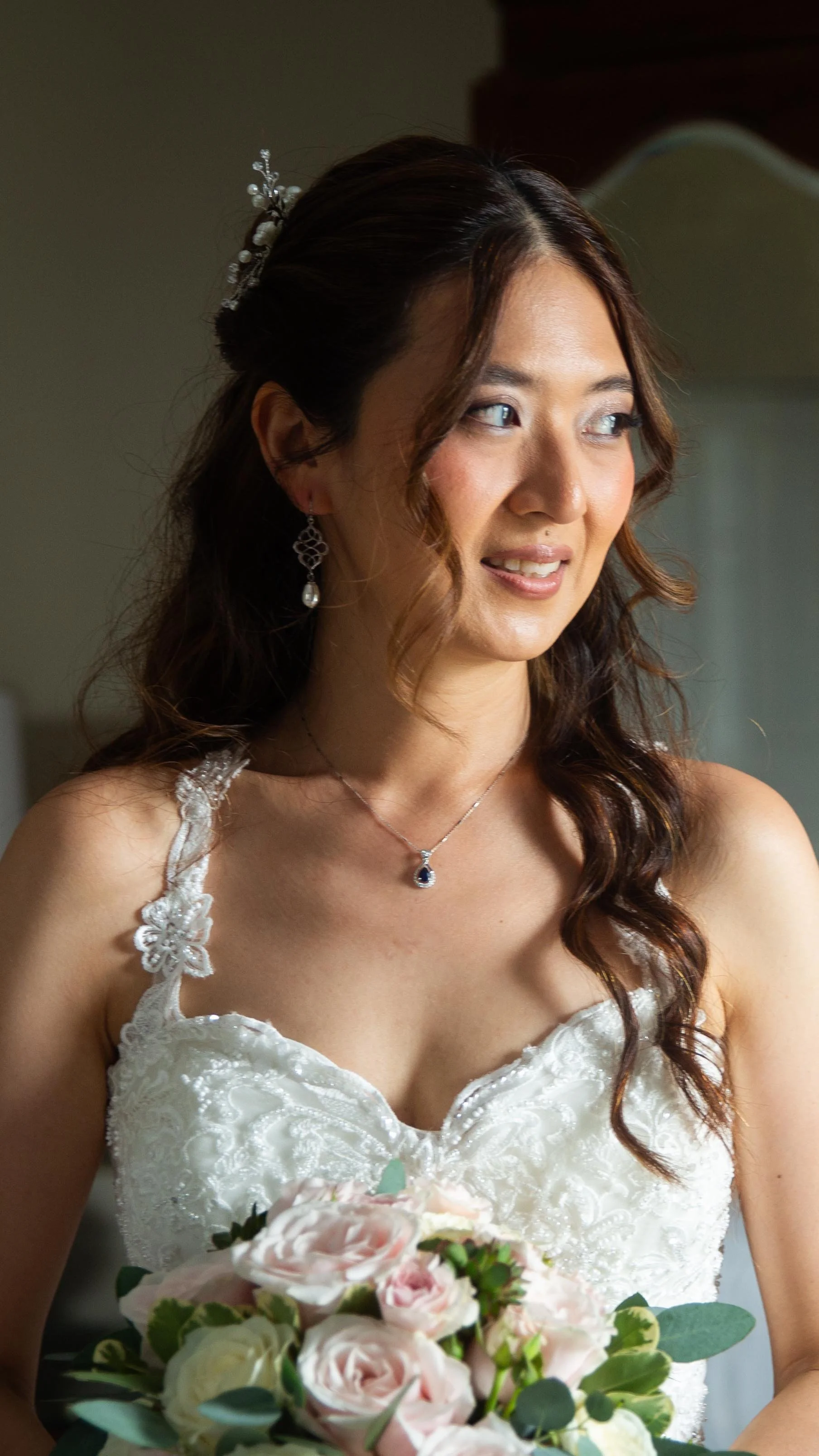 A bride in a white lace wedding dress holding a bouquet of pale pink and white roses, with long wavy brown hair and jewelry, looking to the side indoors.