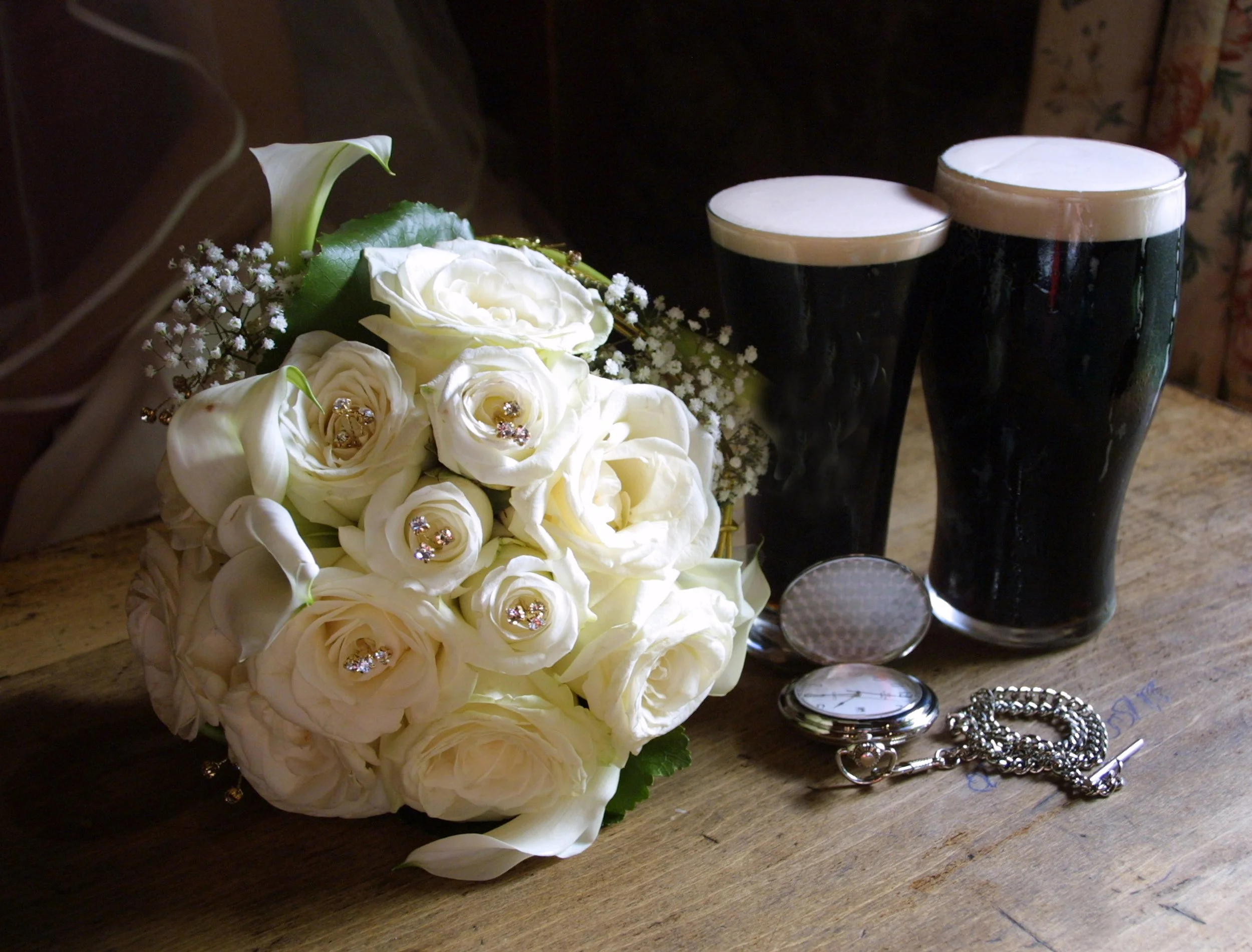 A wedding bouquet of white roses and calla lilies decorated with rhinestones, two glasses of dark stout beer, a pocket watch, a bracelet, and a necklace on a wooden table.