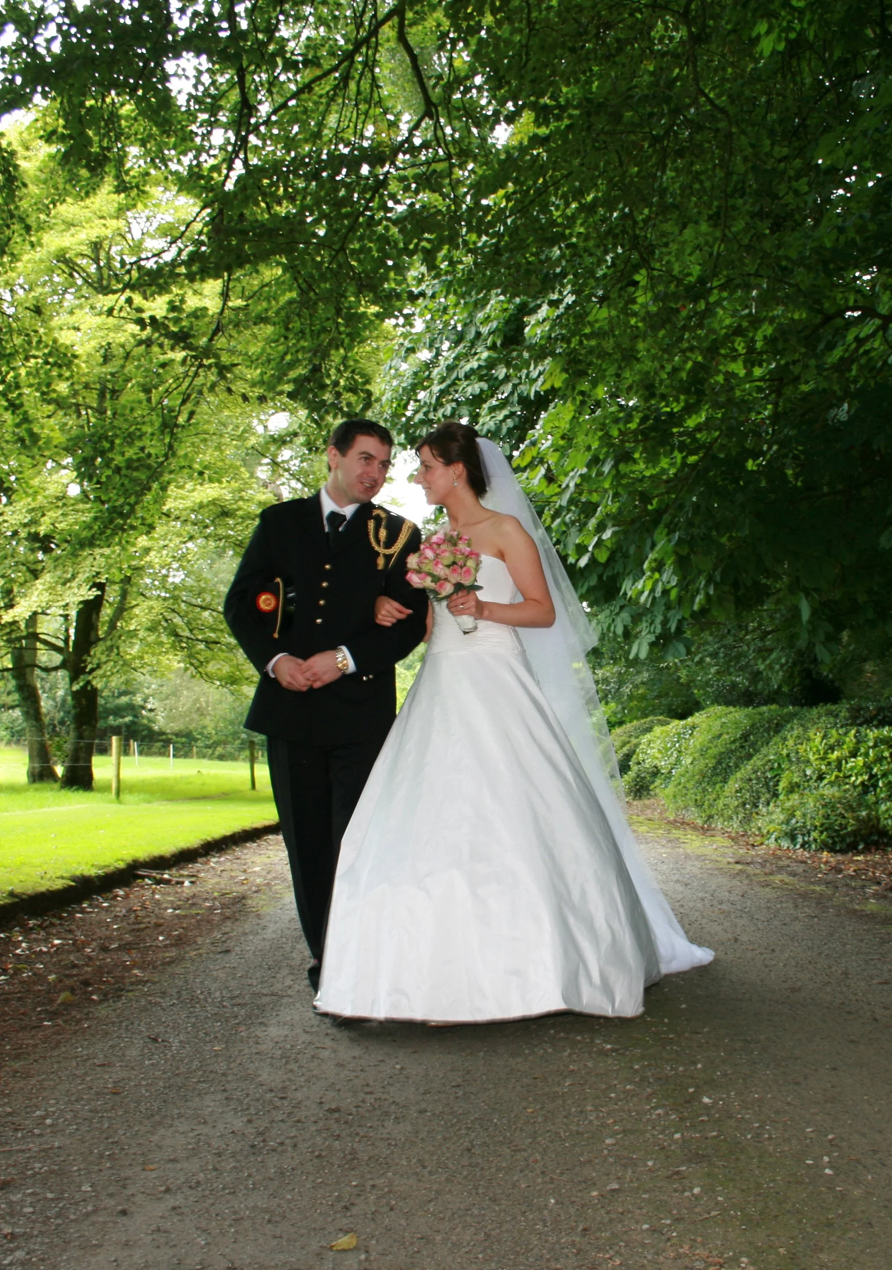 A bride and groom walking together on a wooded path during their wedding day, with the bride holding a bouquet of pink and white flowers.
