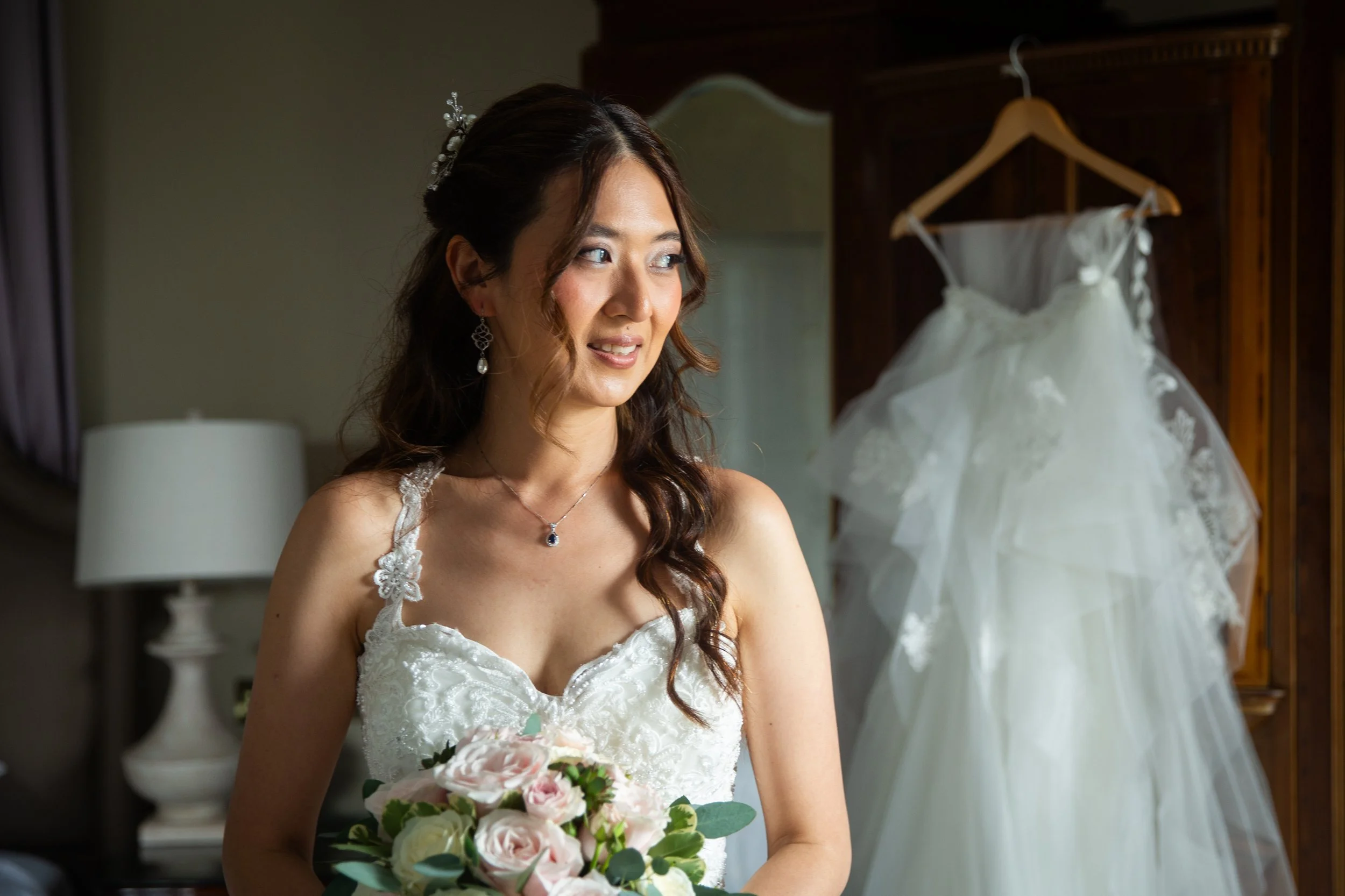 A bride with wavy brown hair standing indoors holding a bouquet of pink and white roses, wearing a white lace wedding dress with floral details and earrings, with a wedding gown hanging on a wooden wardrobe behind her.
