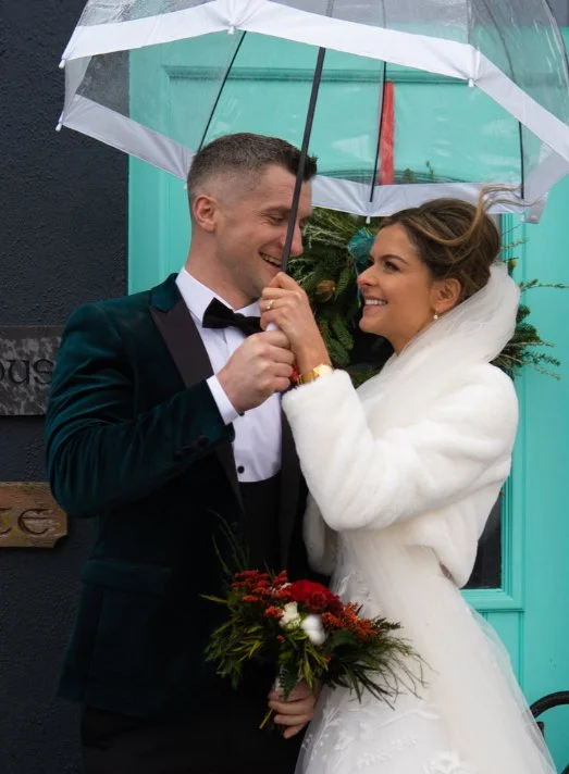 A bride and groom standing outdoors in the rain, smiling at each other under a transparent umbrella. The bride is holding a bouquet of red and white flowers and is wearing a white wedding dress with a fur shawl, while the groom is dressed in a black tuxedo with a bow tie.
