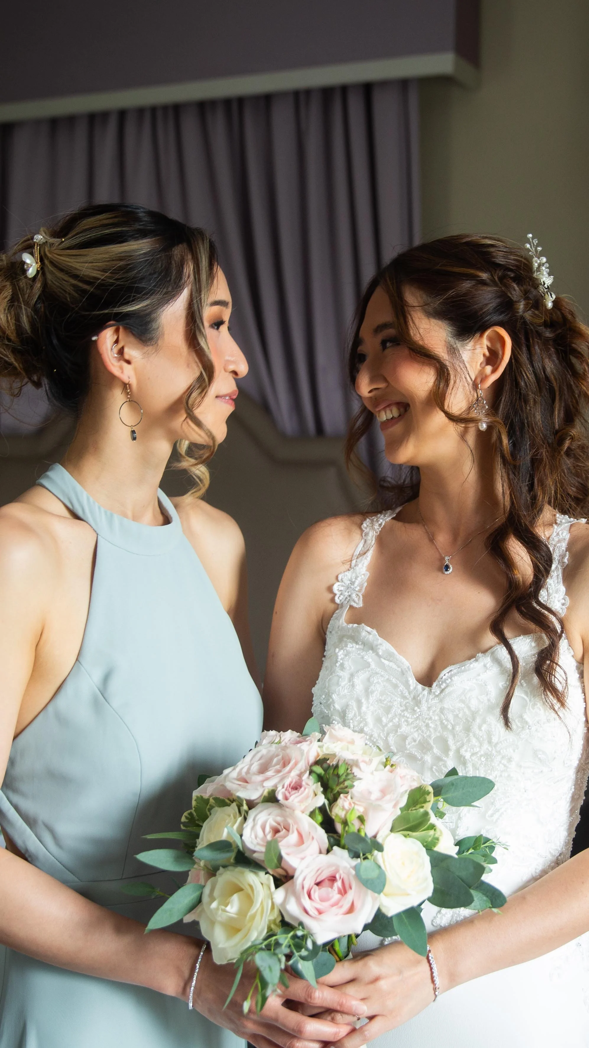 Two women, one in a light blue dress and the other in a white wedding dress, smiling at each other and holding a bouquet of pink and white roses in a room with purple curtains.