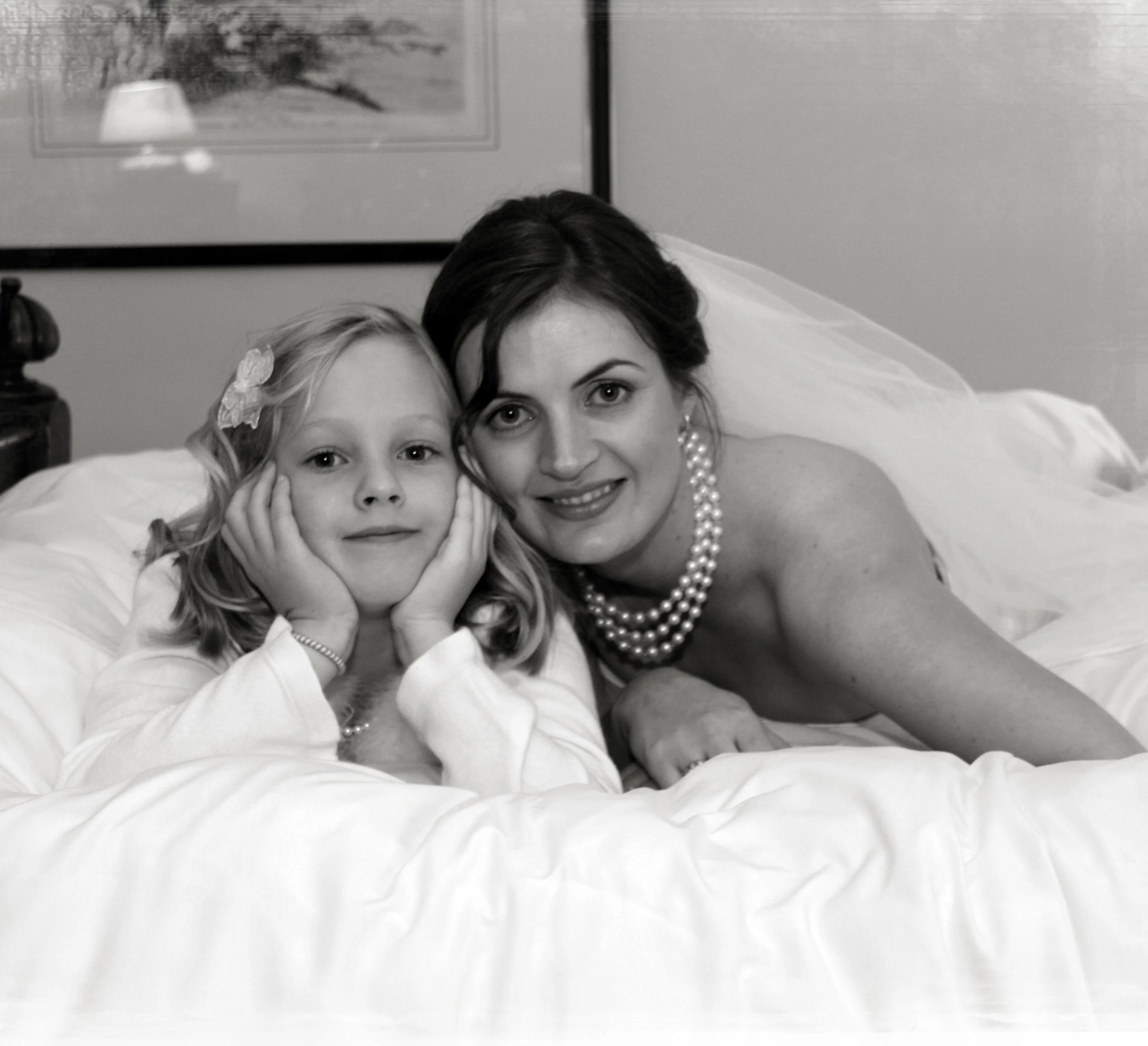 Black and white photograph of a bride and her niece lying on a bed. The bride is wearing a pearl necklace, and the girl has a flower hair clip. They are resting on their elbows, close together, looking at the camera.
