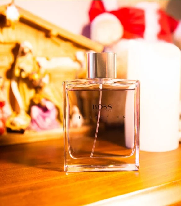 A bottle of BOSS perfume on a wooden surface with a blurred background including Christmas decorations like a Santa figure and a red stocking.