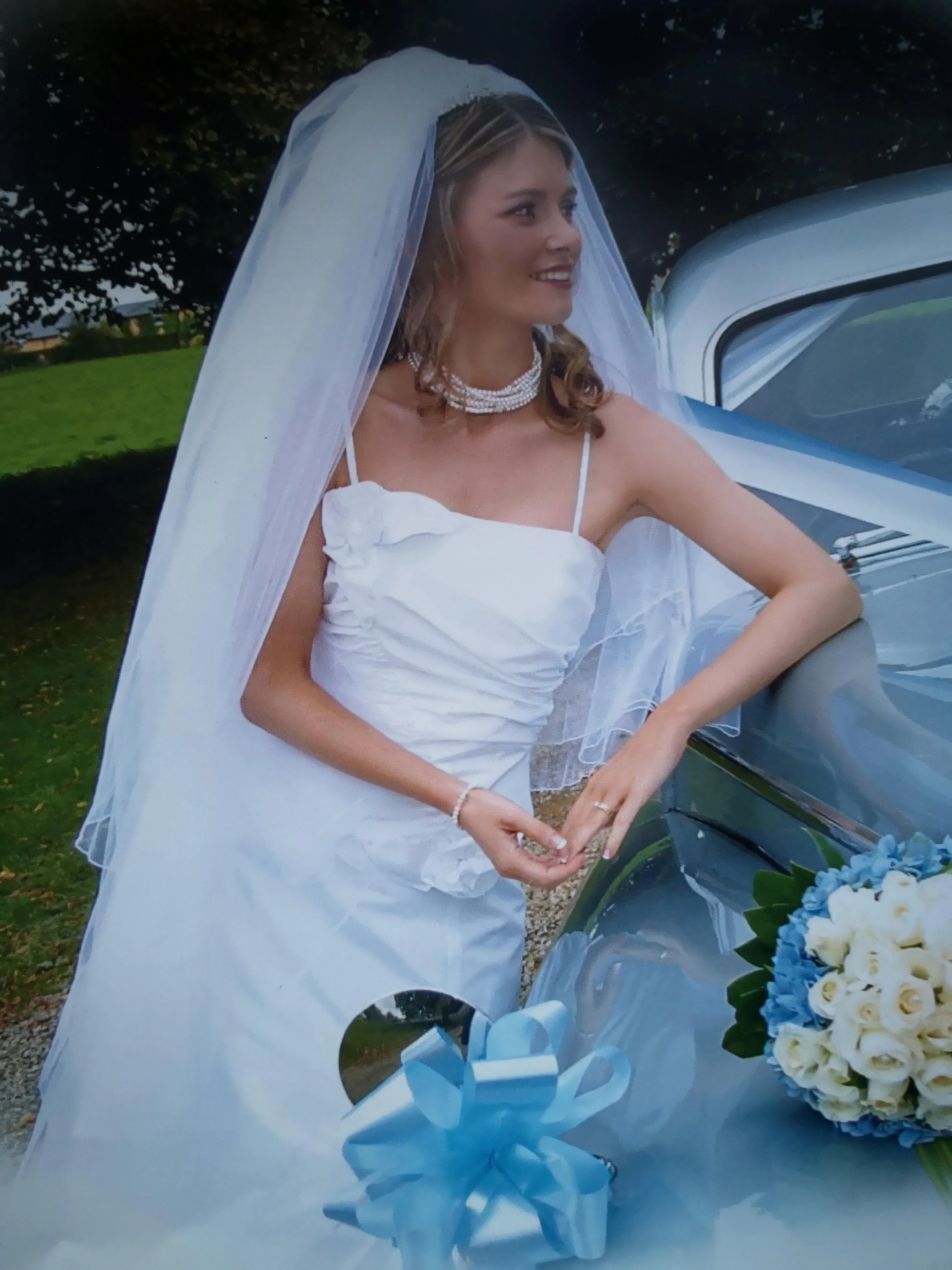 A bride in a white wedding dress with a veil, standing next to a vintage car, holding a bouquet of white roses and blue flowers.