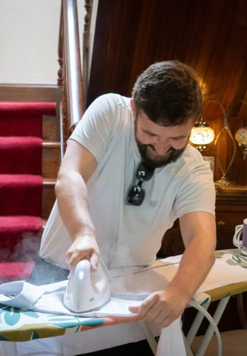 Man with sunglasses hanging from shirt collar and beard ironing clothes on an ironing board in a cozy room.