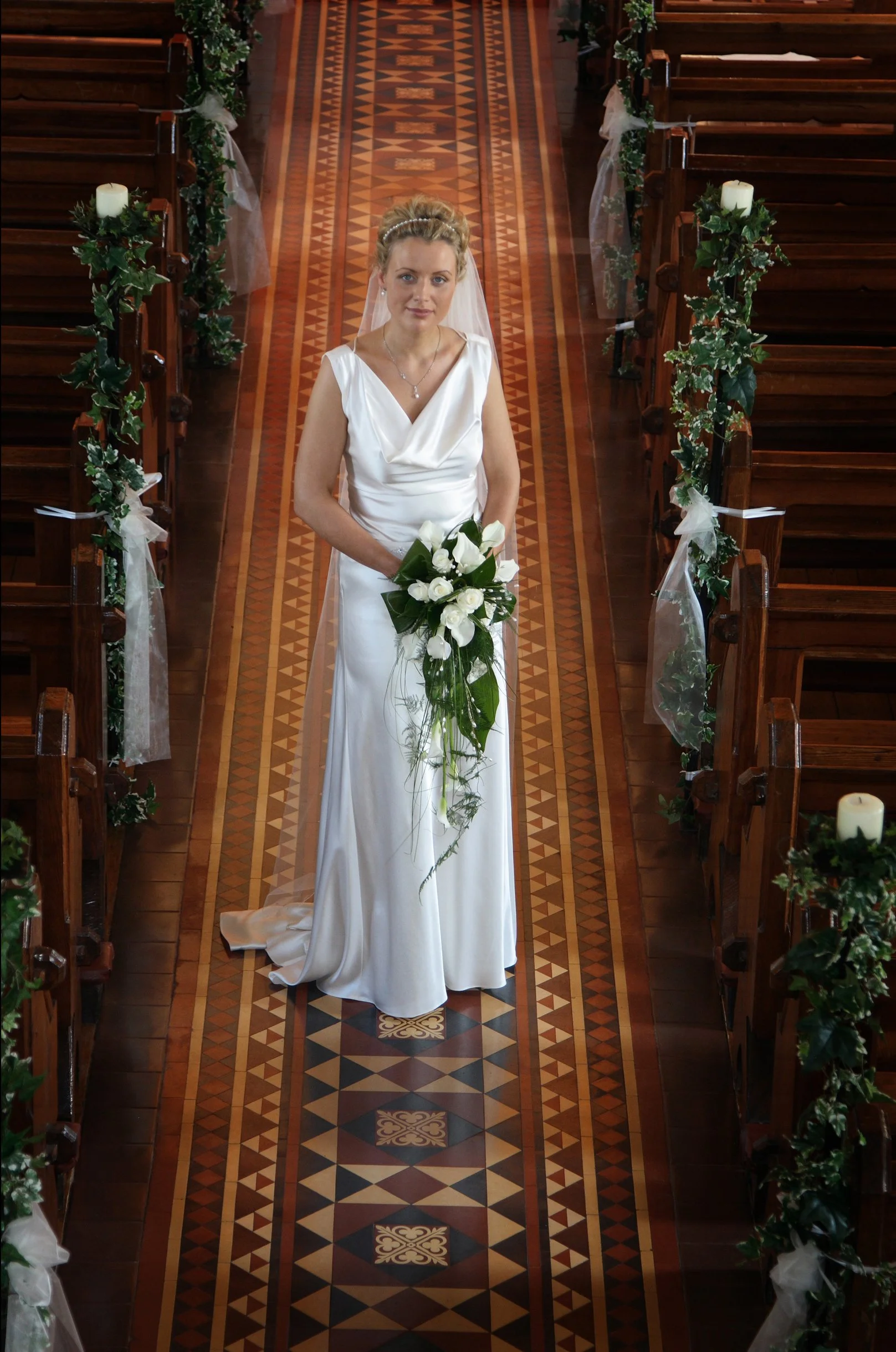 A bride standing in a church aisle, holding a bouquet of white flowers, dressed in a white satin gown with a veil and styled hair, surrounded by wooden pews decorated with green and white floral arrangements and candles.