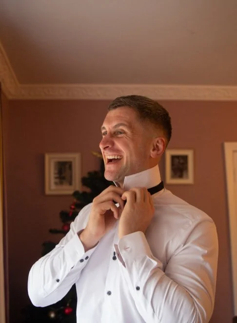 Man in white shirt adjusting bow tie in a warmly decorated room with Christmas tree in the background.