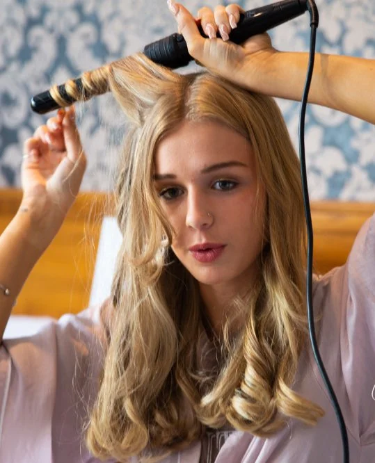 A young woman with blonde hair is curling her hair with a curling iron.