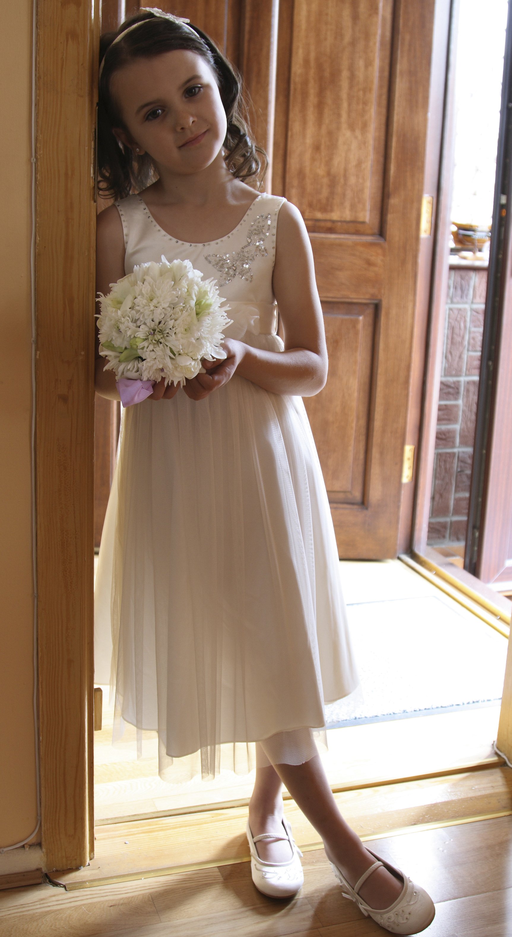 A young flower girl on a wedding day in a white dress holding a bouquet of white flowers, standing in a doorway.