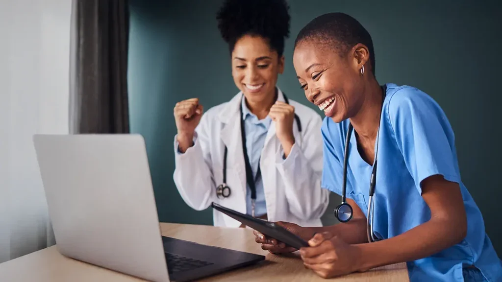 Two female healthcare professionals, one in a white coat and the other in blue scrubs, celebrating together while looking at a tablet in a medical setting.