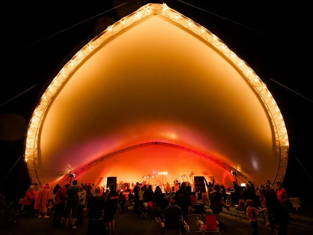 People watching a concert in an outdoor venue with a large, illuminated, teardrop-shaped tent at night.
