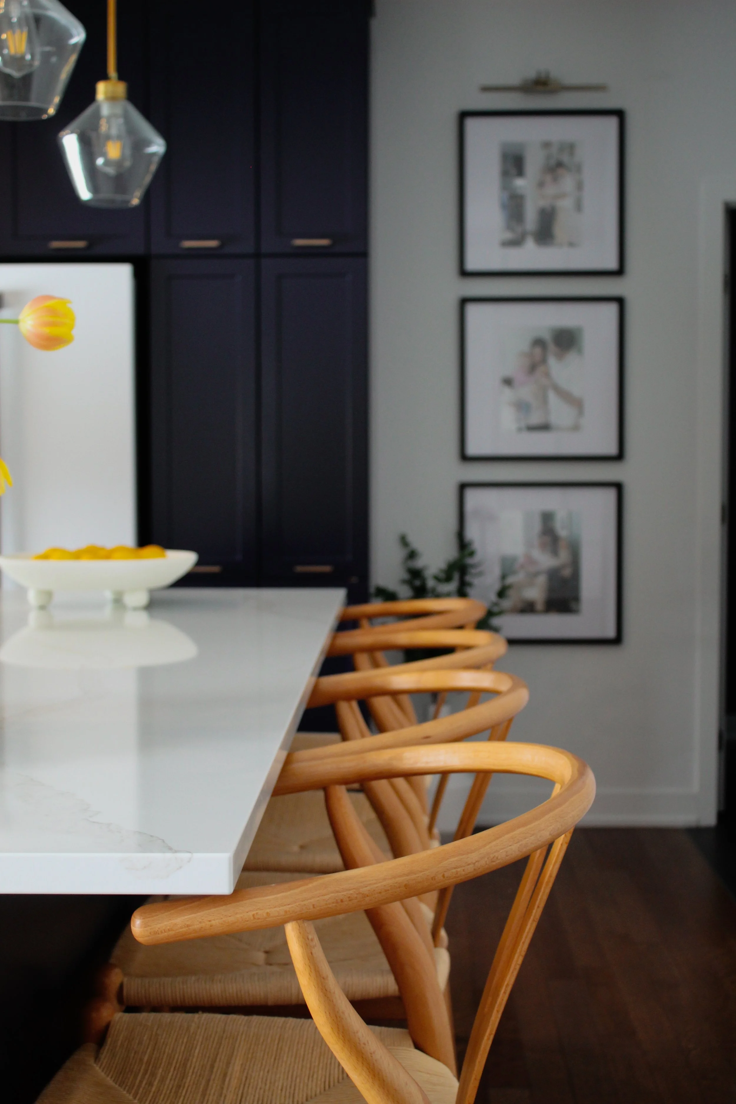 A dining area with a white table and wooden chairs, a black wall cabinet, framed pictures on the wall, and a countertop with a vase of tulips.