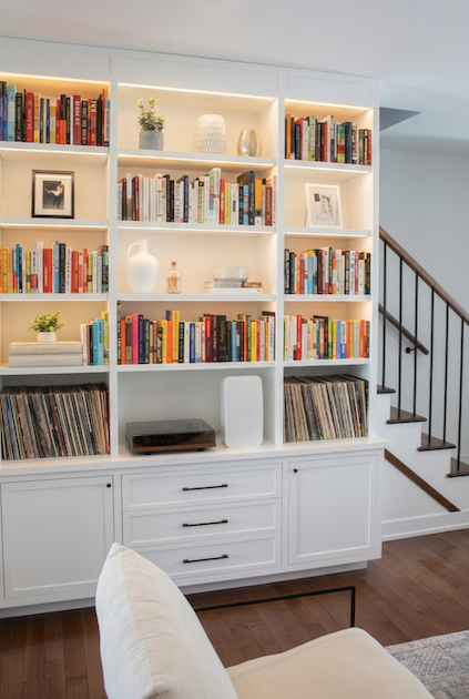 White built-in bookshelf filled with colorful books and decorative items, located in a living room near a staircase.