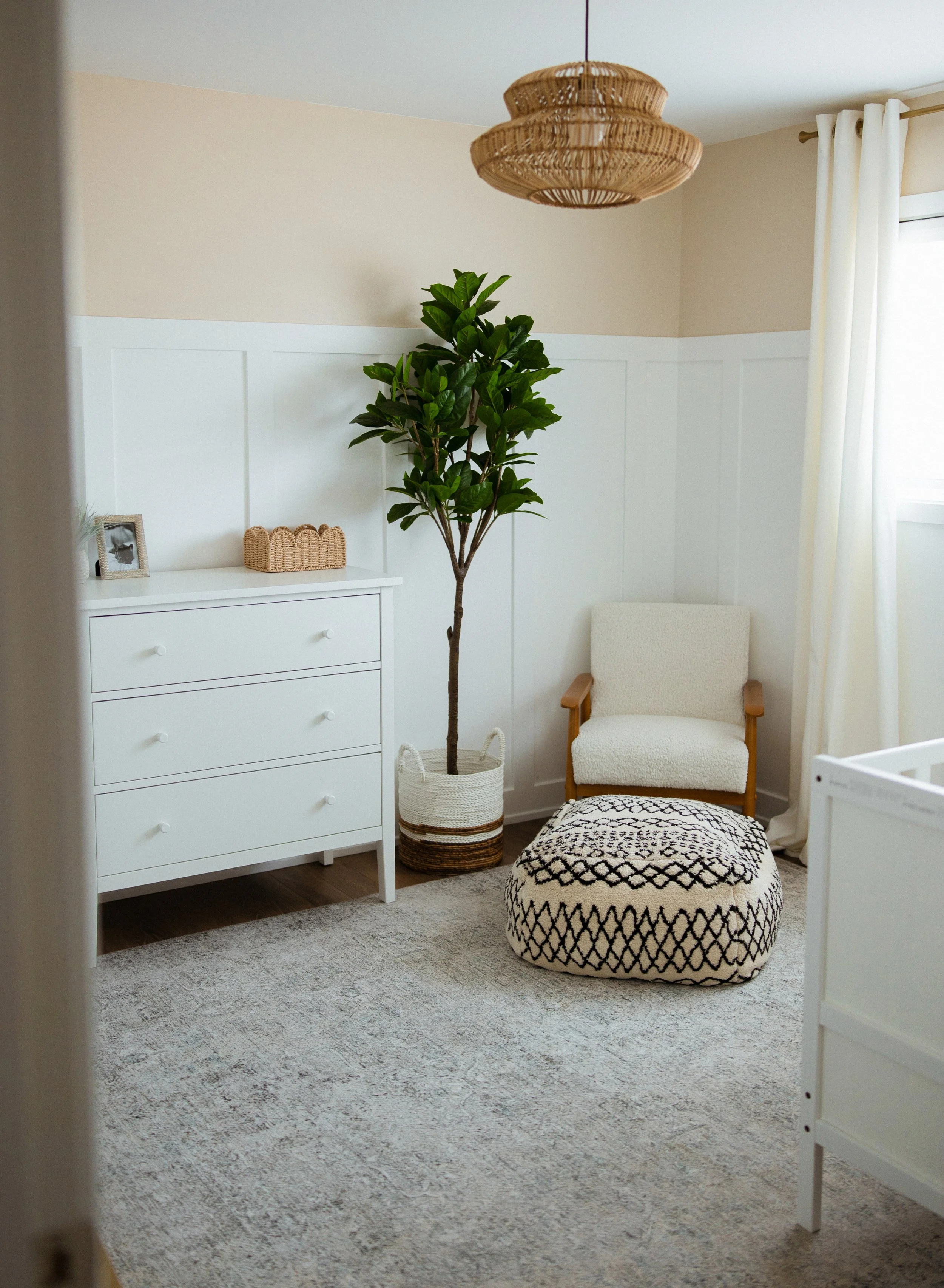A cozy room with a white dresser, a green potted plant, a white armchair, a black and white patterned pouf, a woven basket, a woven ceiling light, and white curtains.