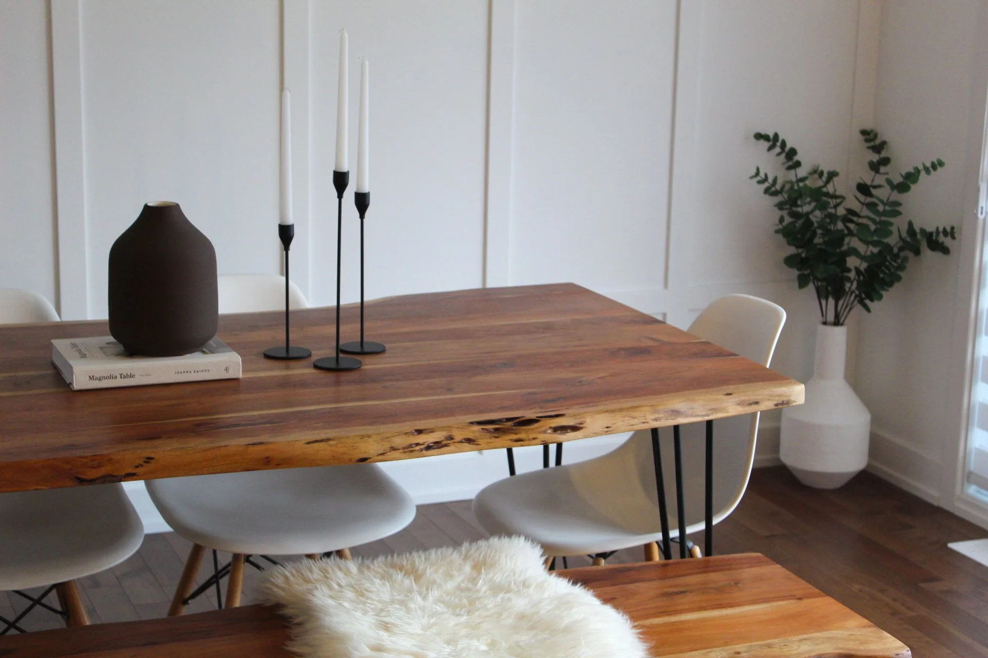 A wooden dining table with a dark brown vase and three white candlestick holders. The table is surrounded by white chairs, and a fur throw is draped over one of the chairs. In the background, a potted plant is visible in the corner of the room, again