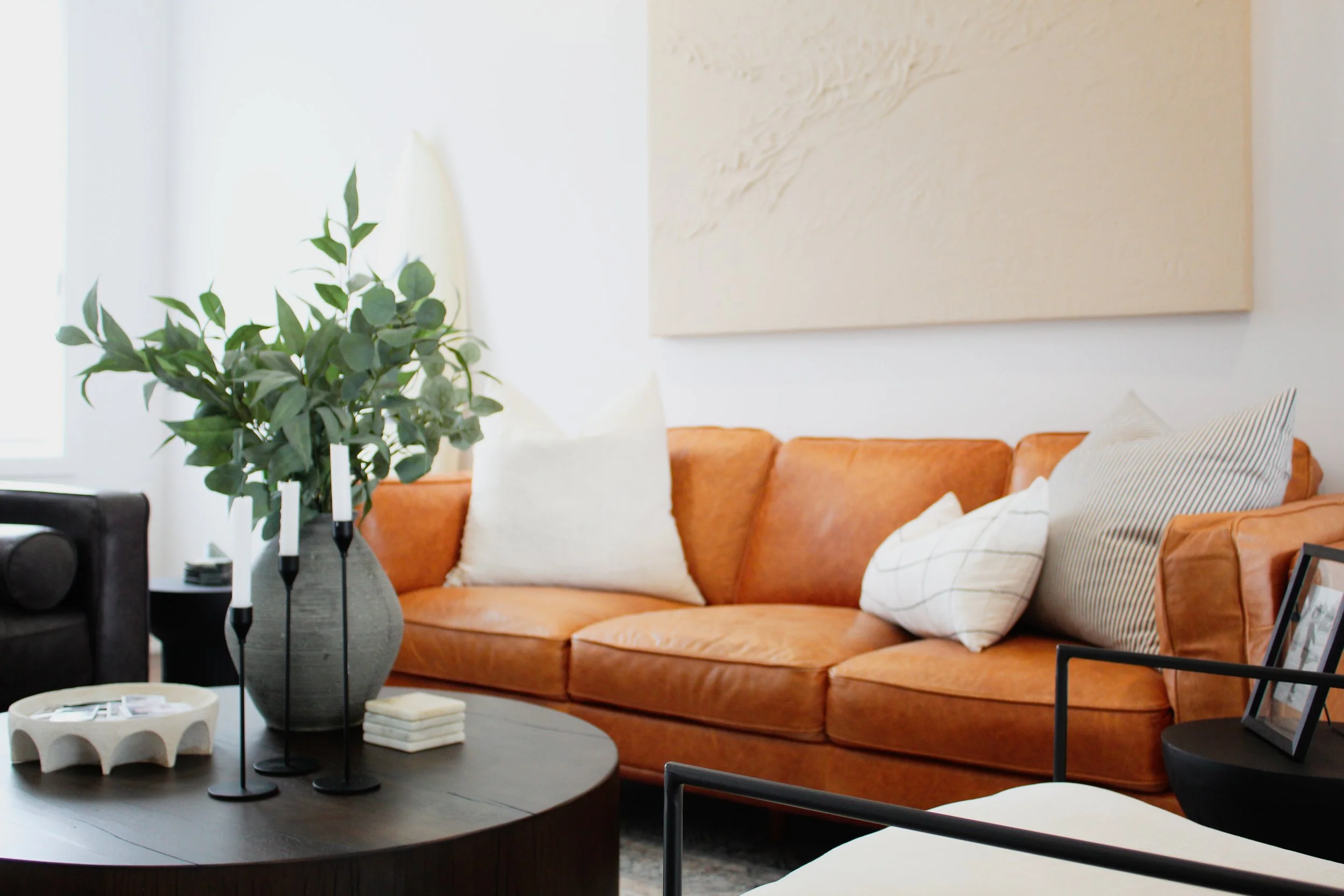 Living room with an orange sofa, white pillows, a large abstract painting, a black side table, a potted plant, and decorative items on a round coffee table.