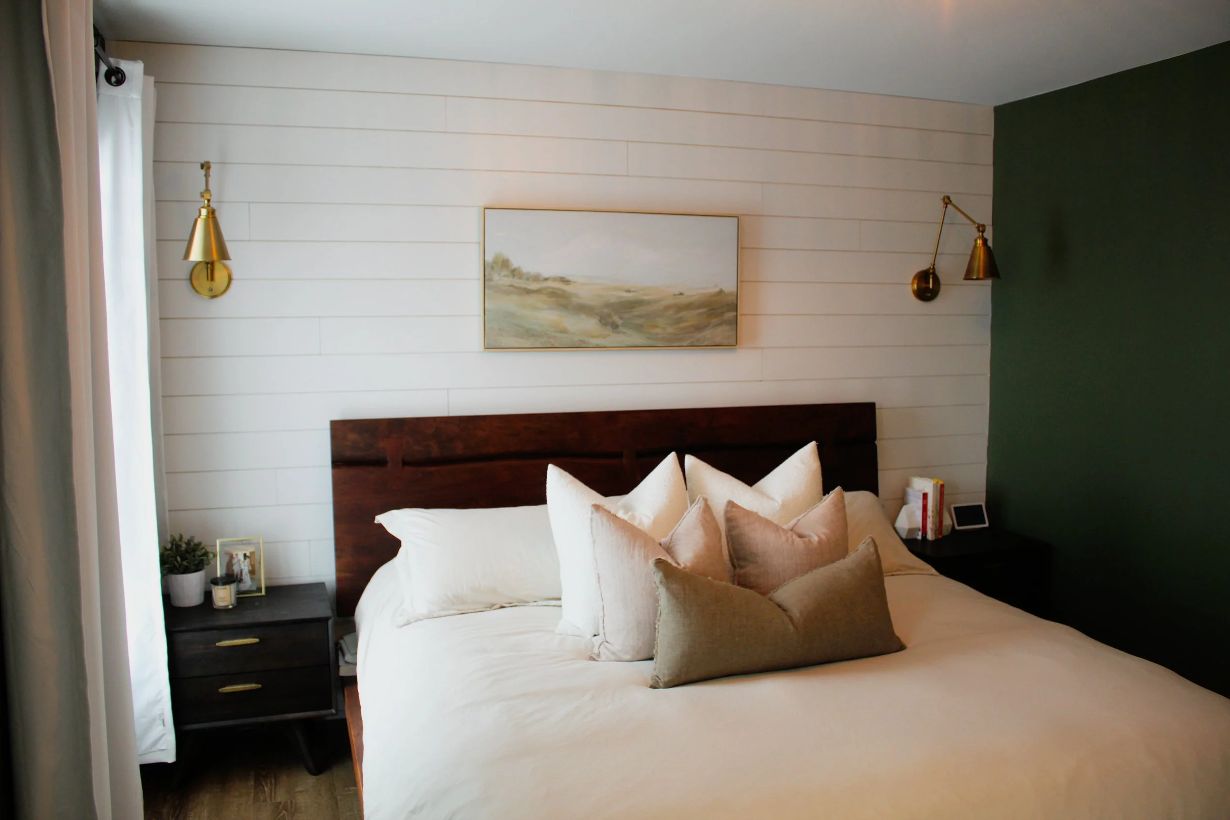 A bedroom with white wooden paneled walls, a large bed with white and beige pillows, two gold wall-mounted lamps, a dark wooden headboard, and a nightstand with a framed photo, a candle, and a small plant. There is a green accent wall and a painting 