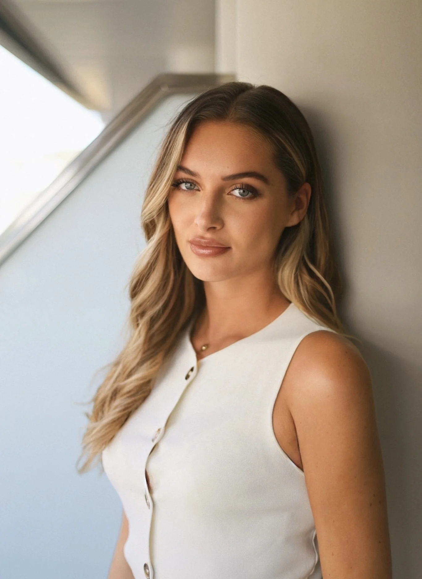 A young woman with long wavy blonde hair, blue eyes, and a subtle makeup look, wearing a sleeveless white top, standing indoors against a light-colored wall.