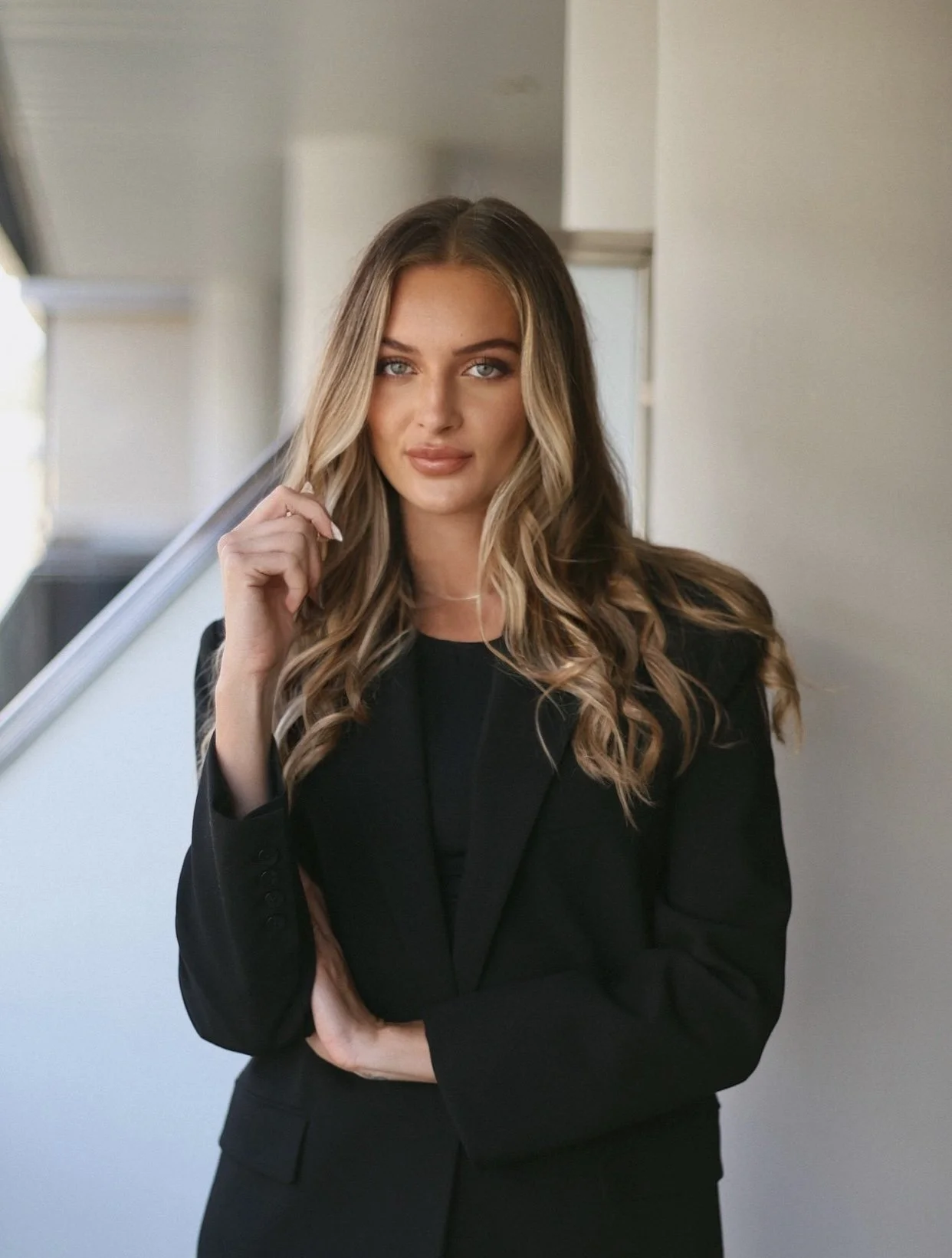 A young woman with light brown, wavy hair and blue eyes, wearing a black blazer, standing indoors with a blurred background of stairs and walls.