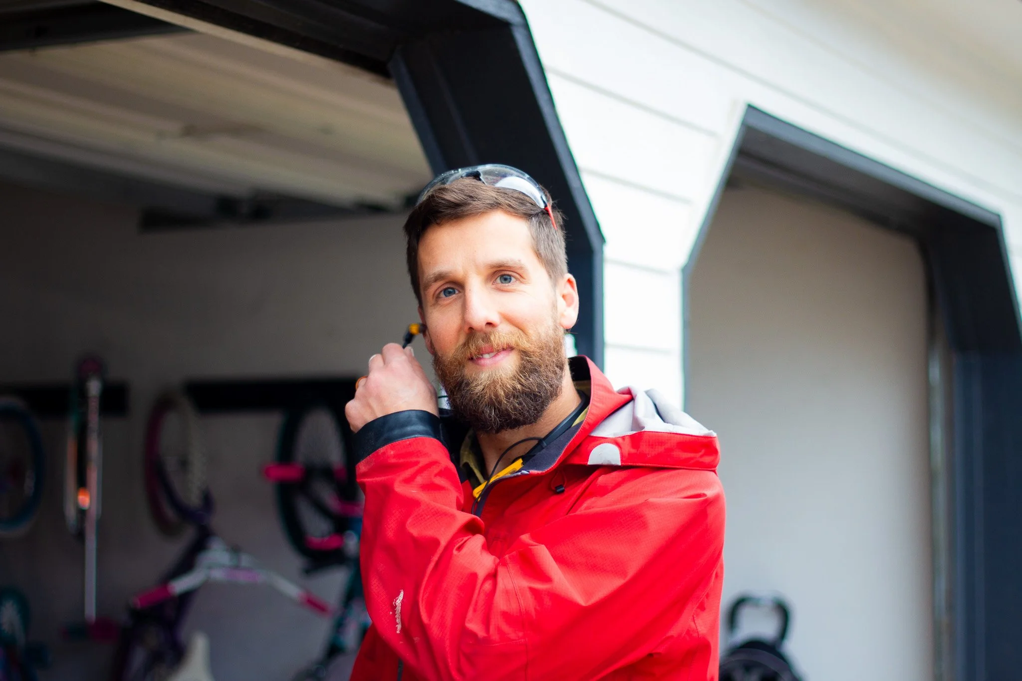 A man with a beard and blue eyes wearing a red jacket and safety glasses on his head is standing in a garage or workshop, adjusting his ear protection. Bicycles are hanging in the background.
