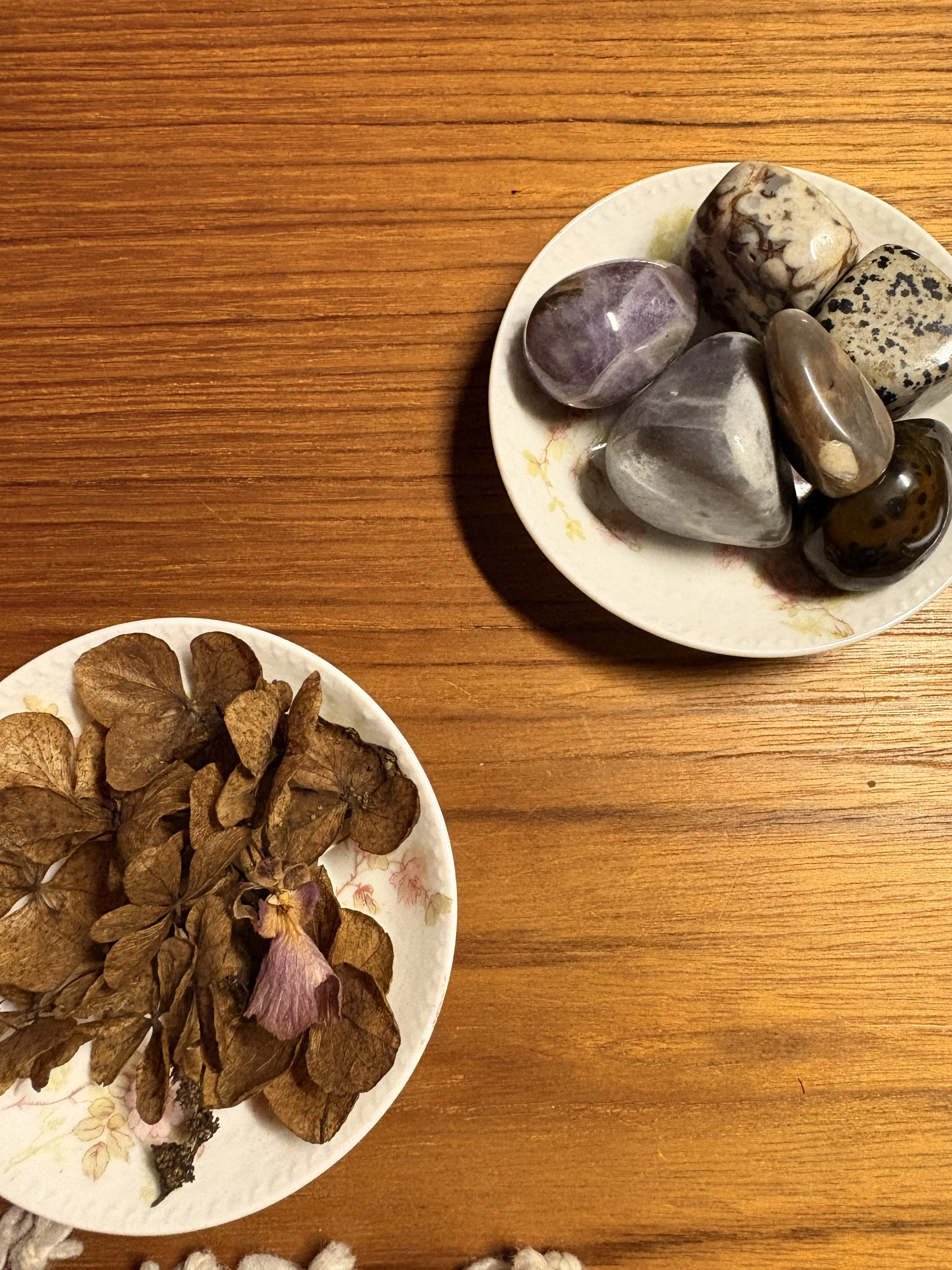 Dried hydrangea flowers on a floral-patterned plate and a collection of assorted polished stones on a white floral plate, both on a wooden surface.