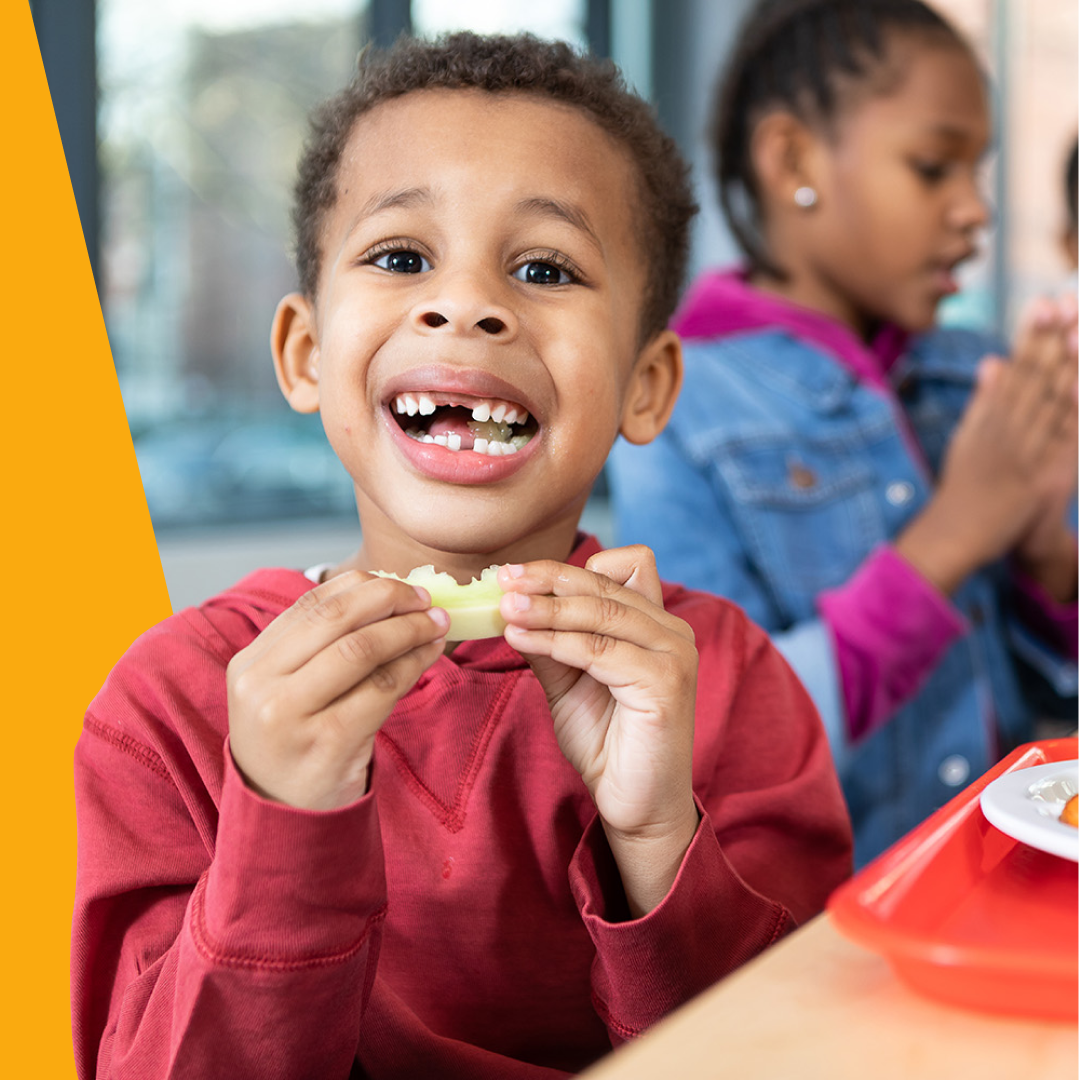 A young boy with a missing front tooth smiling while eating a snack, with a girl in the background focused on her meal.