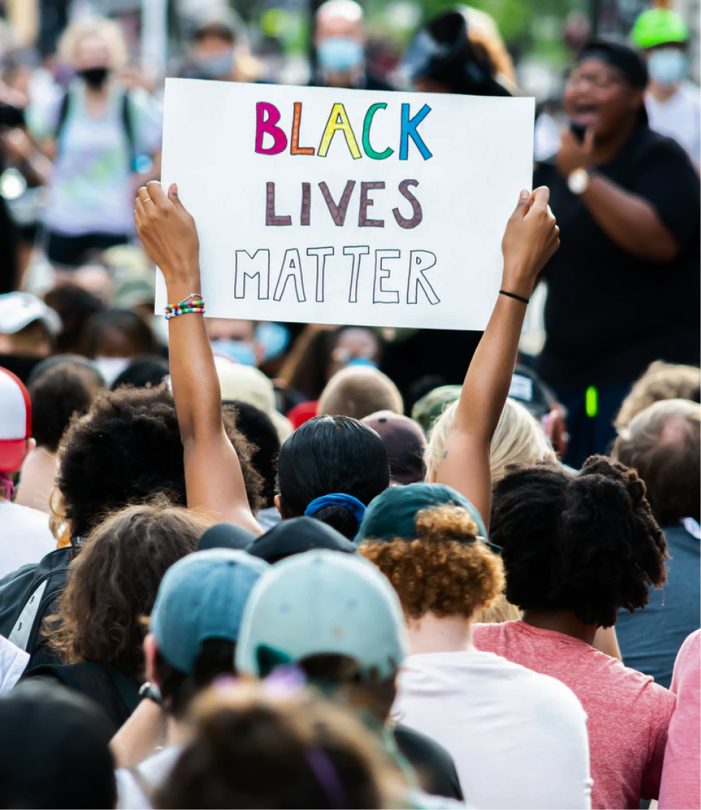 A person in a crowd at a protest holding a white sign that says 'Black Lives Matter' in colorful letters.