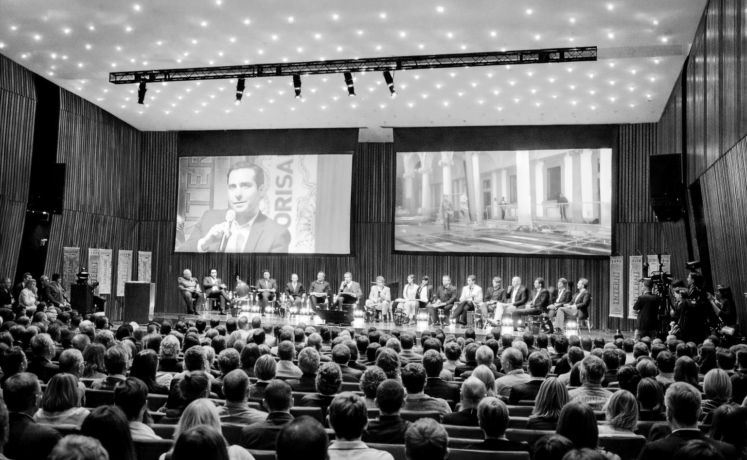 A large auditorium filled with a seated audience watching a panel discussion on stage, with numerous participants seated in chairs, two large screens above displaying close ups of speakers, and professional camera crews capturing the event.