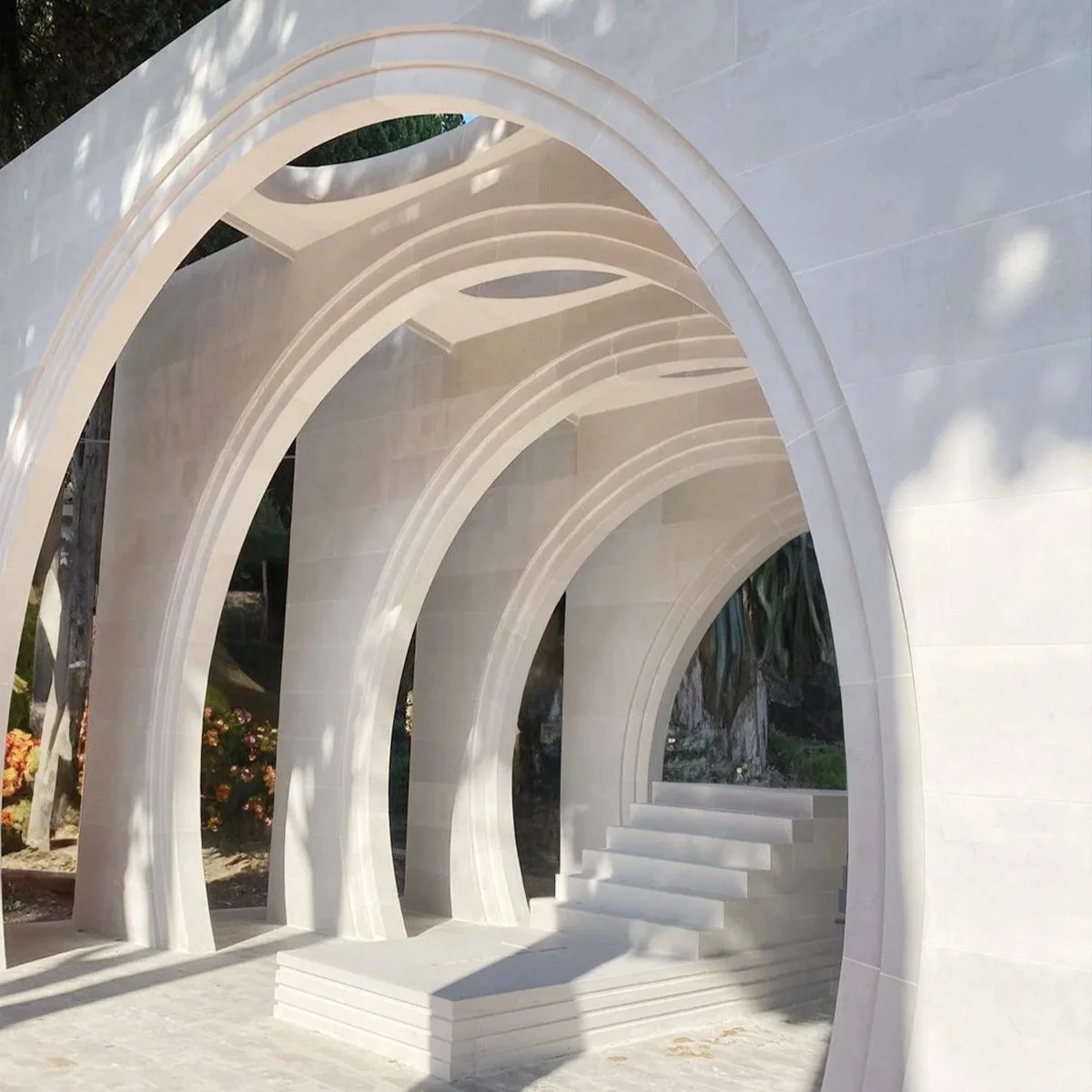 Arch-shaped white marble structure with steps and open spaces, casting shadows on the ground.