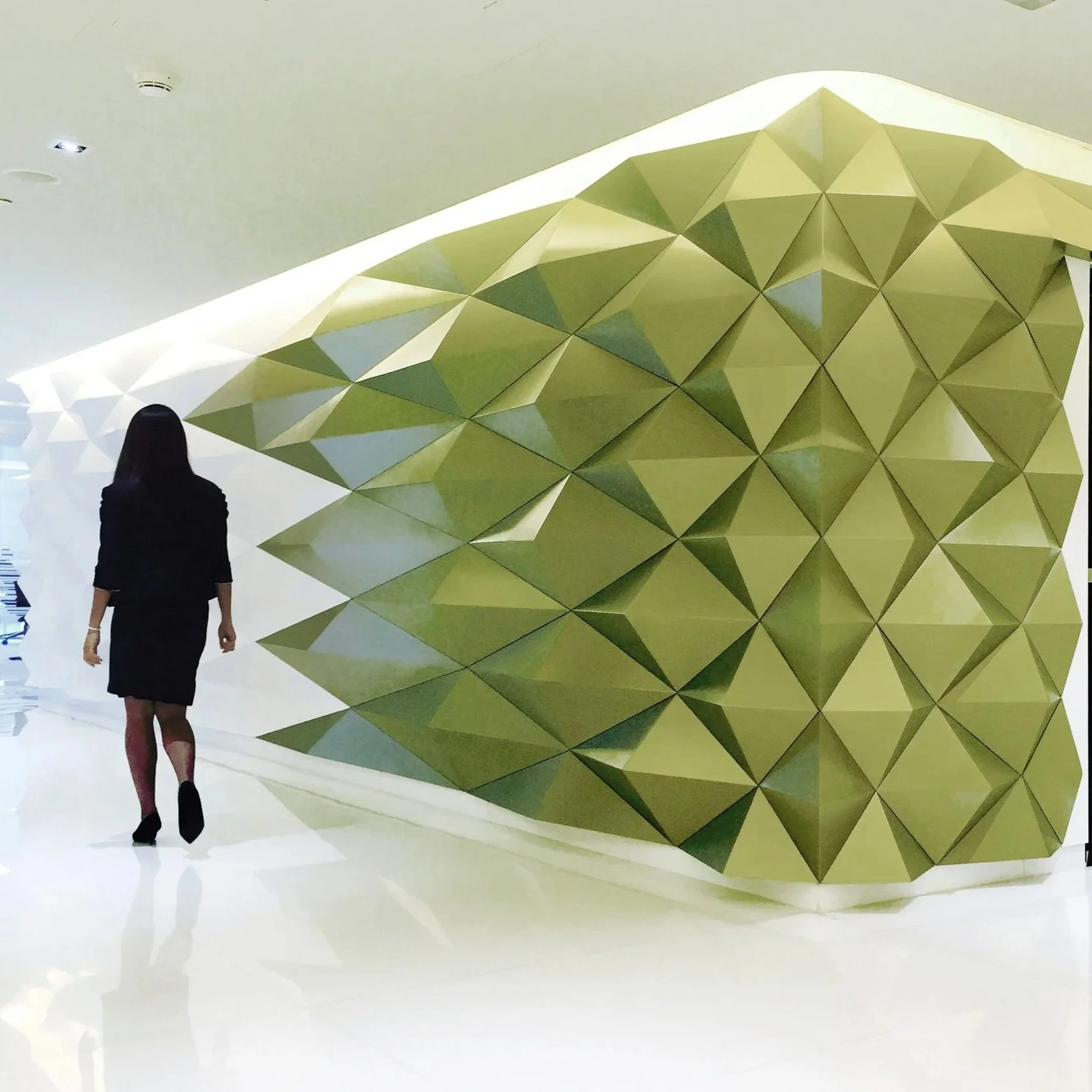 A woman dressed in black walking past a geometric yellow-green sculpture in a modern, minimalistic indoor space.