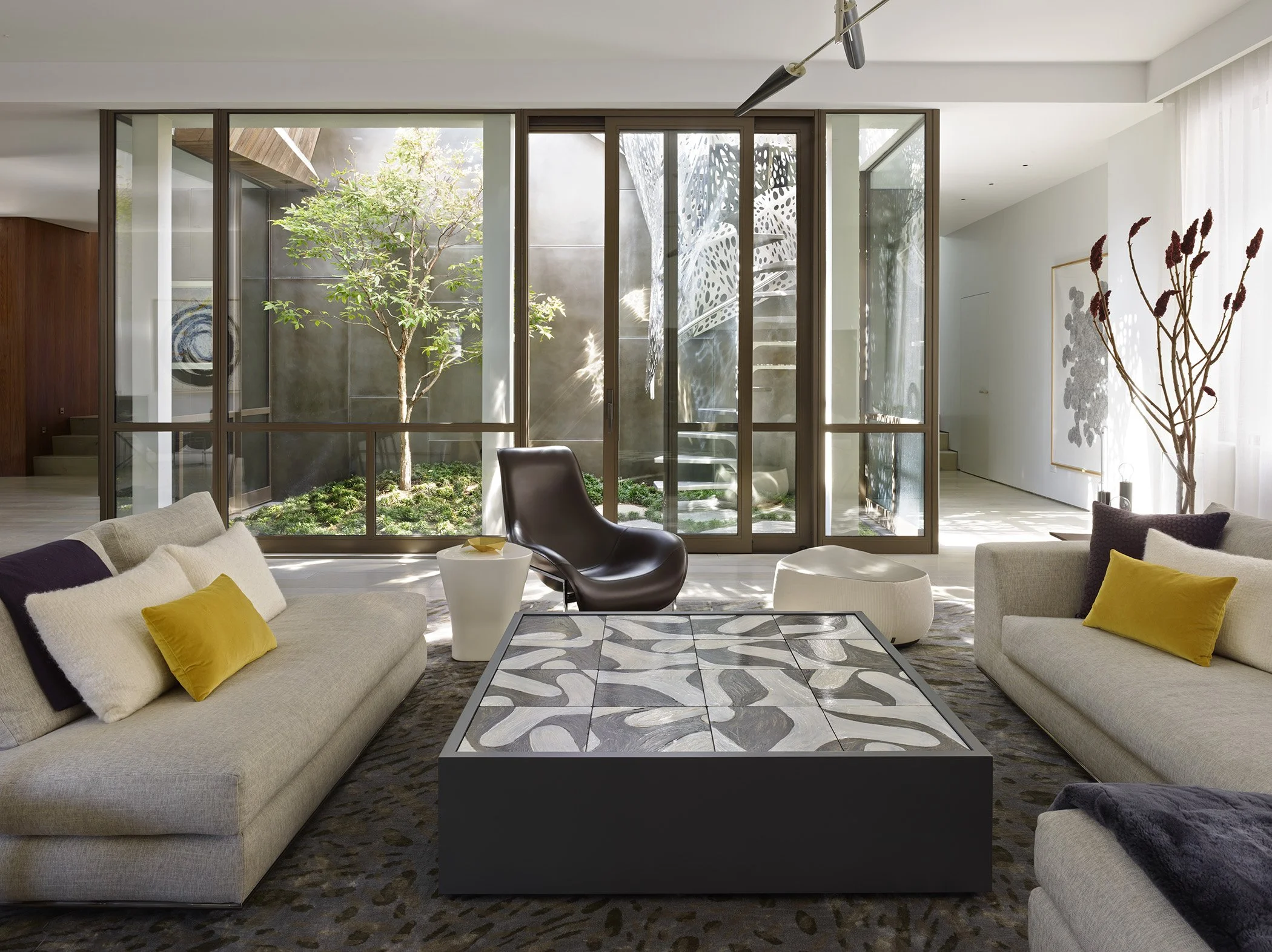 Modern living room with large glass windows facing a small outdoor garden with a tree, contemporary furniture including two beige sofas with pillows, a black leather chair, a patterned coffee table, and decorative vases with branches, bright natural lighting.